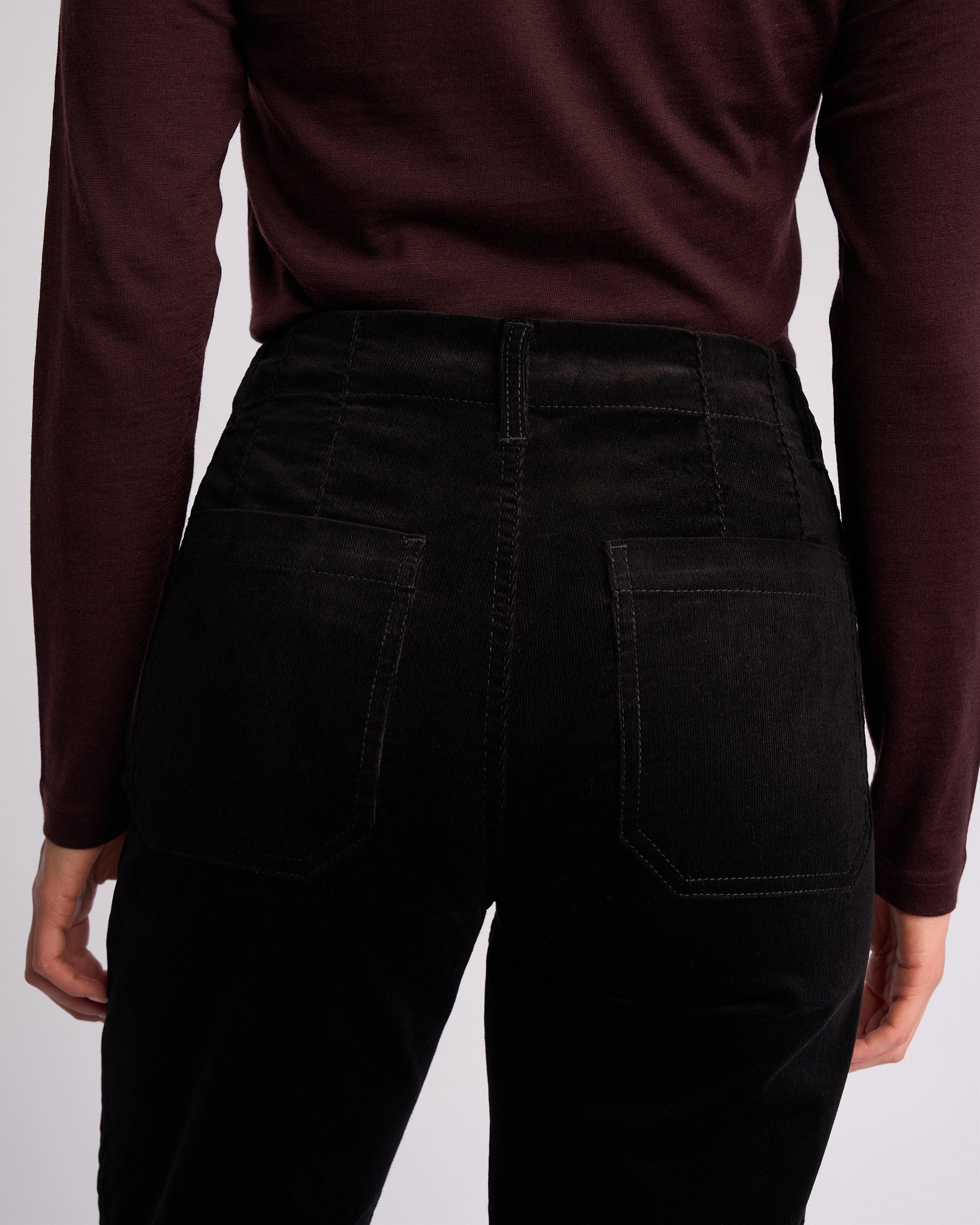 A person is shown from the back, wearing a long-sleeved maroon top and Vassalli Daily Cord Pant in black, with focus on the waist and hip area.