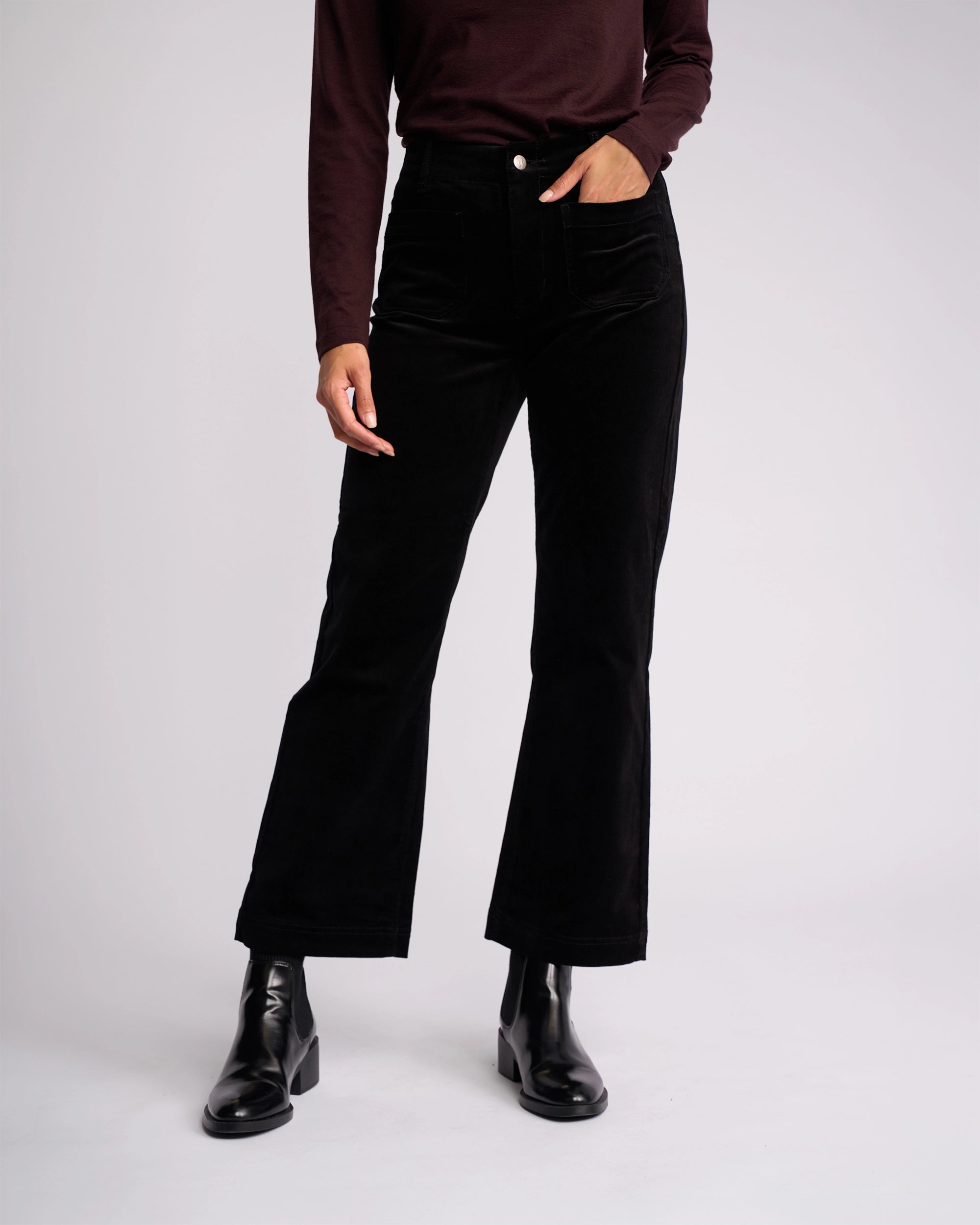 Wearing Vassalli’s Daily Cord Pant in Black, styled with a long-sleeve dark top and black ankle boots, a person stands against a plain light background, their face out of view.