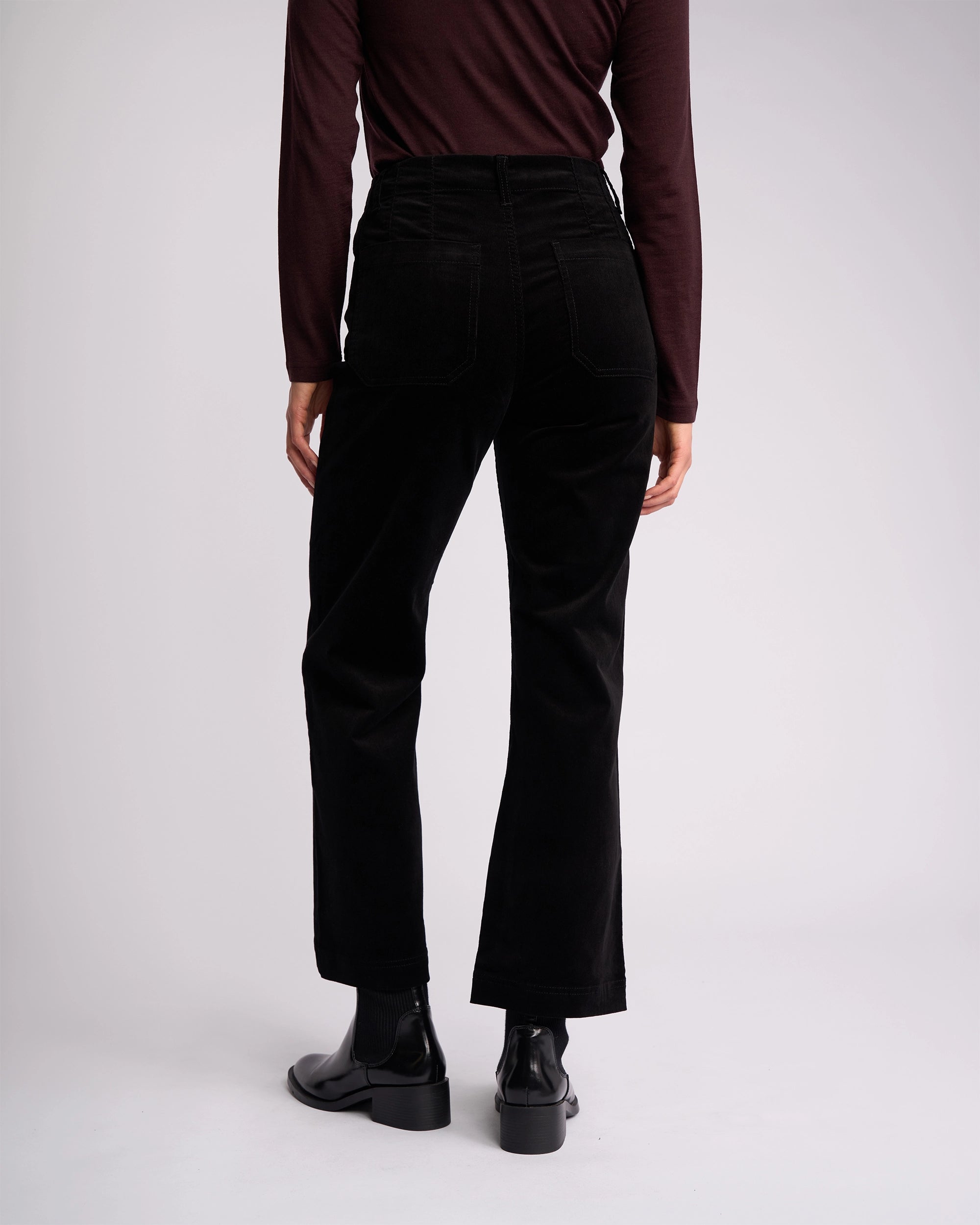 A person faces away from the camera in a long-sleeve dark brown top, Vassalli Daily Cord Pant in black, and black ankle boots against a plain light background.