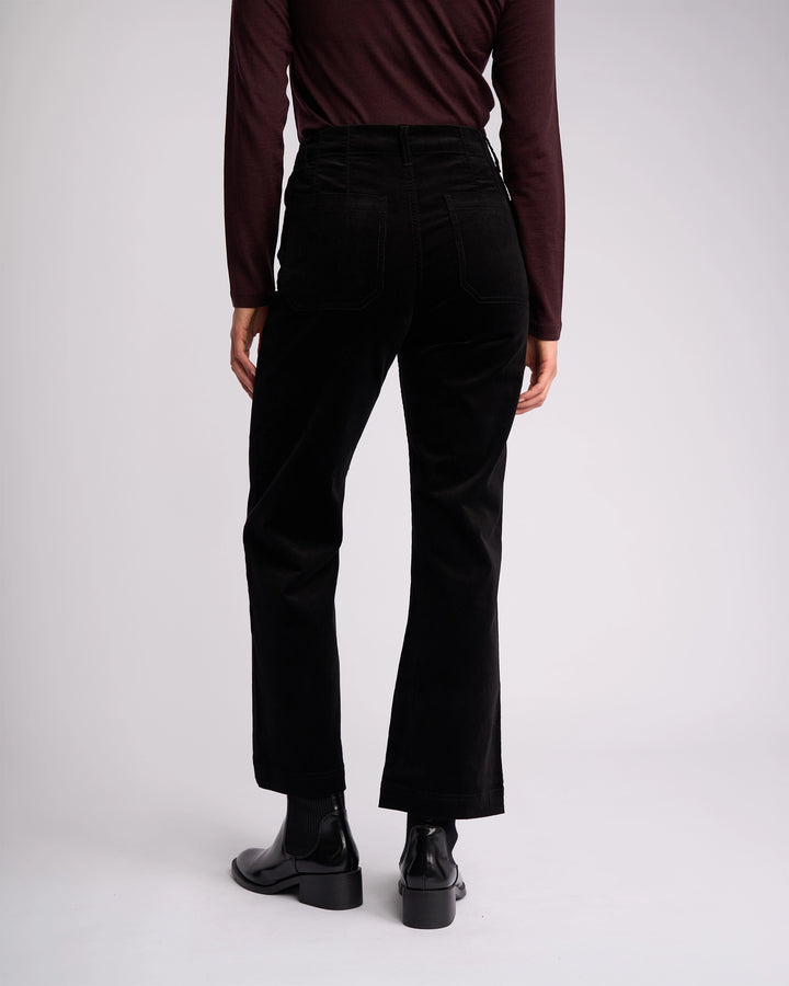A person faces away from the camera in a long-sleeve dark brown top, Vassalli Daily Cord Pant in black, and black ankle boots against a plain light background.