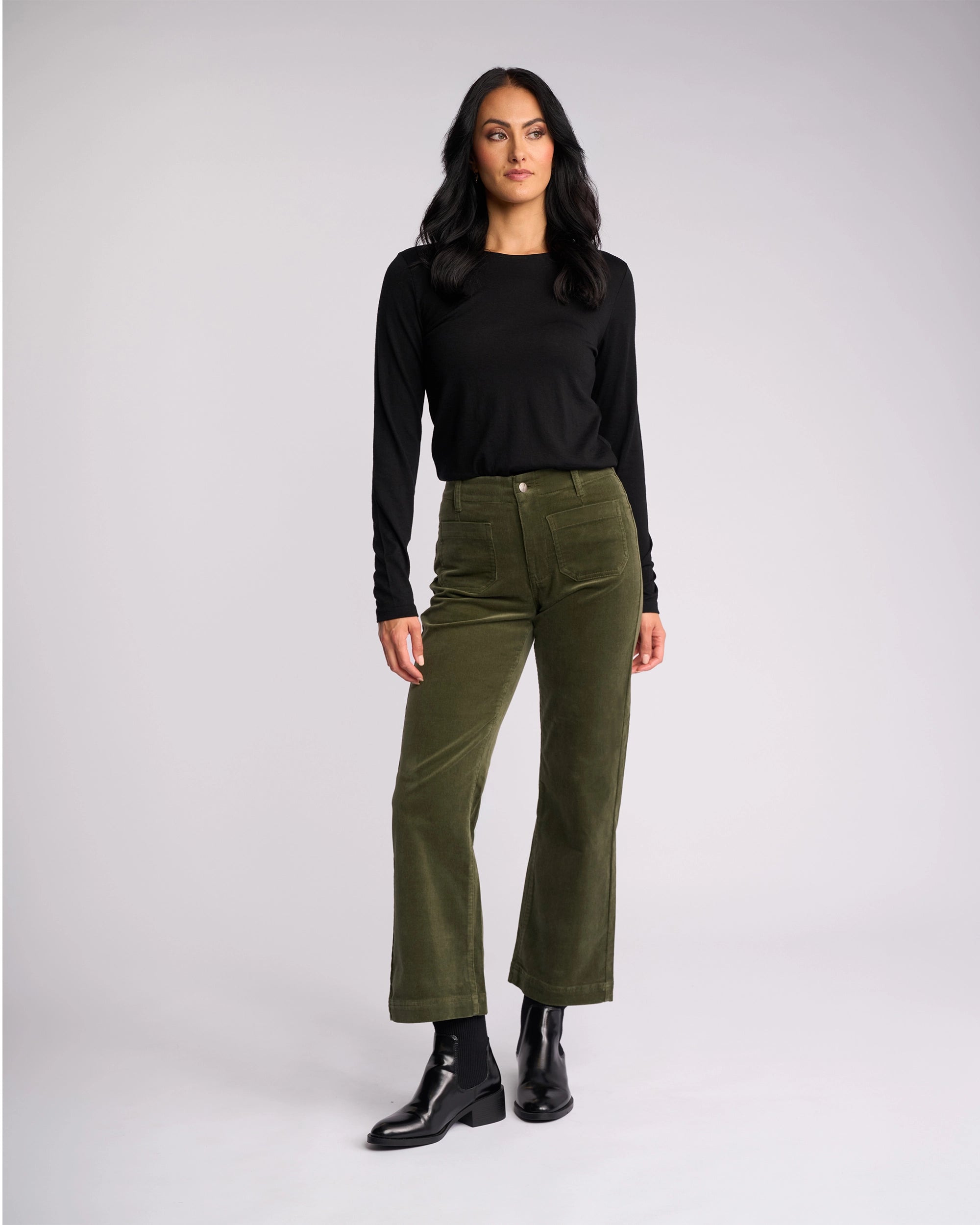 A woman with long dark hair wears Vassalli’s Daily Cord Pant in Deep Sage, a black long-sleeve top, and black ankle boots, standing against a plain light gray background.