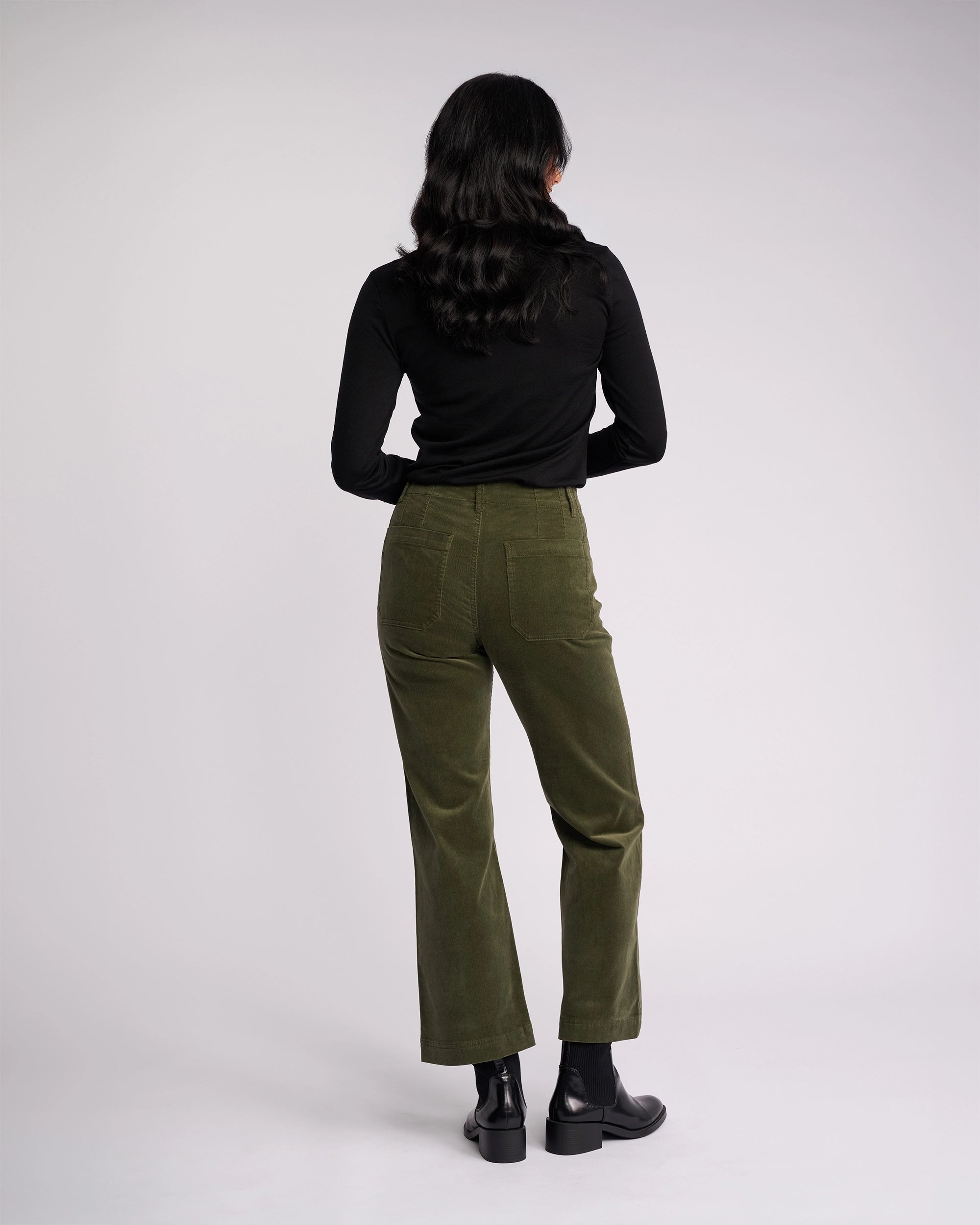 A person with long dark hair, wearing a black long-sleeve top, Vassalli Daily Cord Pant in Deep Sage, and black boots stands facing away against a plain light background.