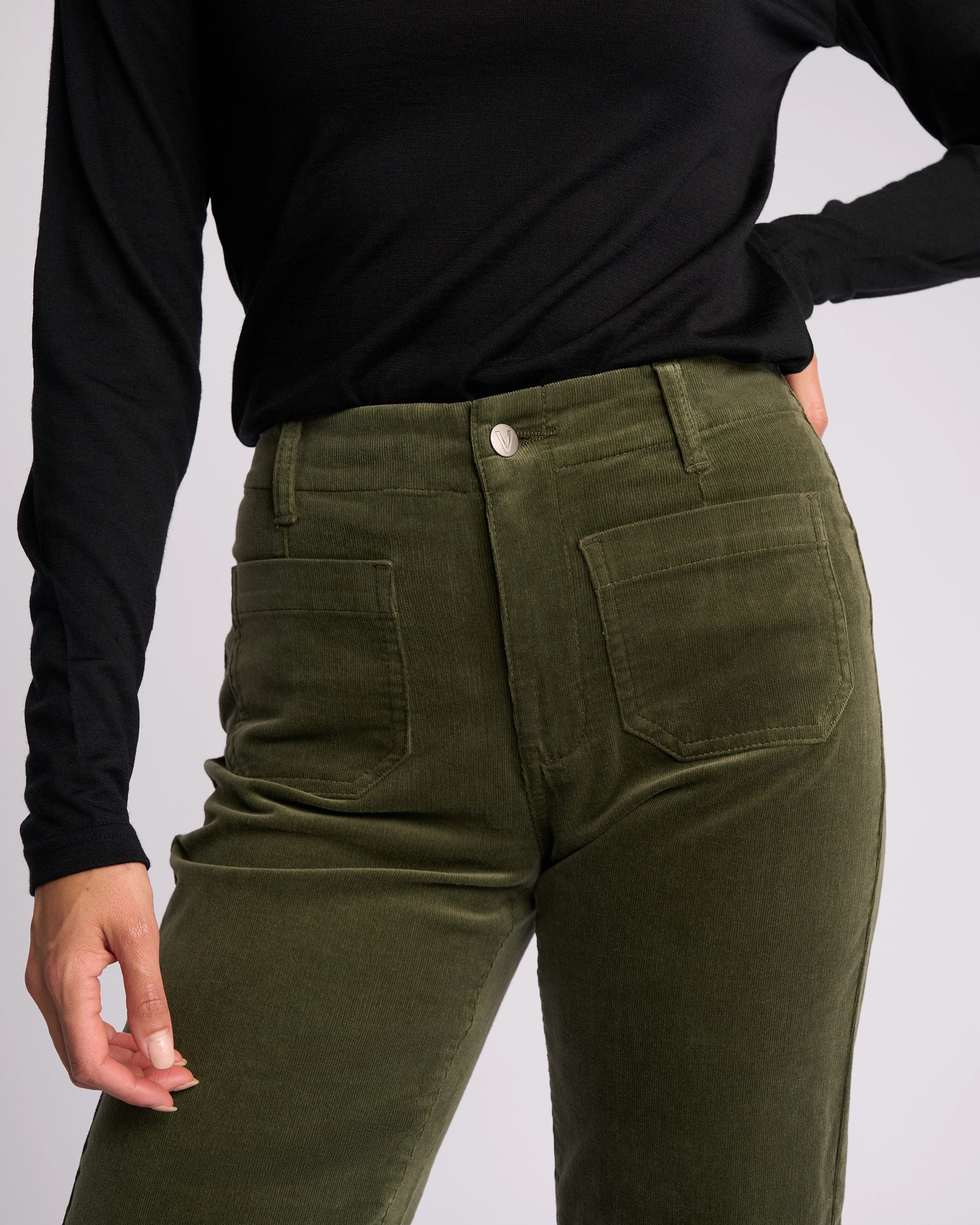 A person wears a black long-sleeve top and Vassalli Daily Cord Pant in Deep Sage, featuring a high-waisted, straight-leg fit with front pockets, standing with one hand on their hip.