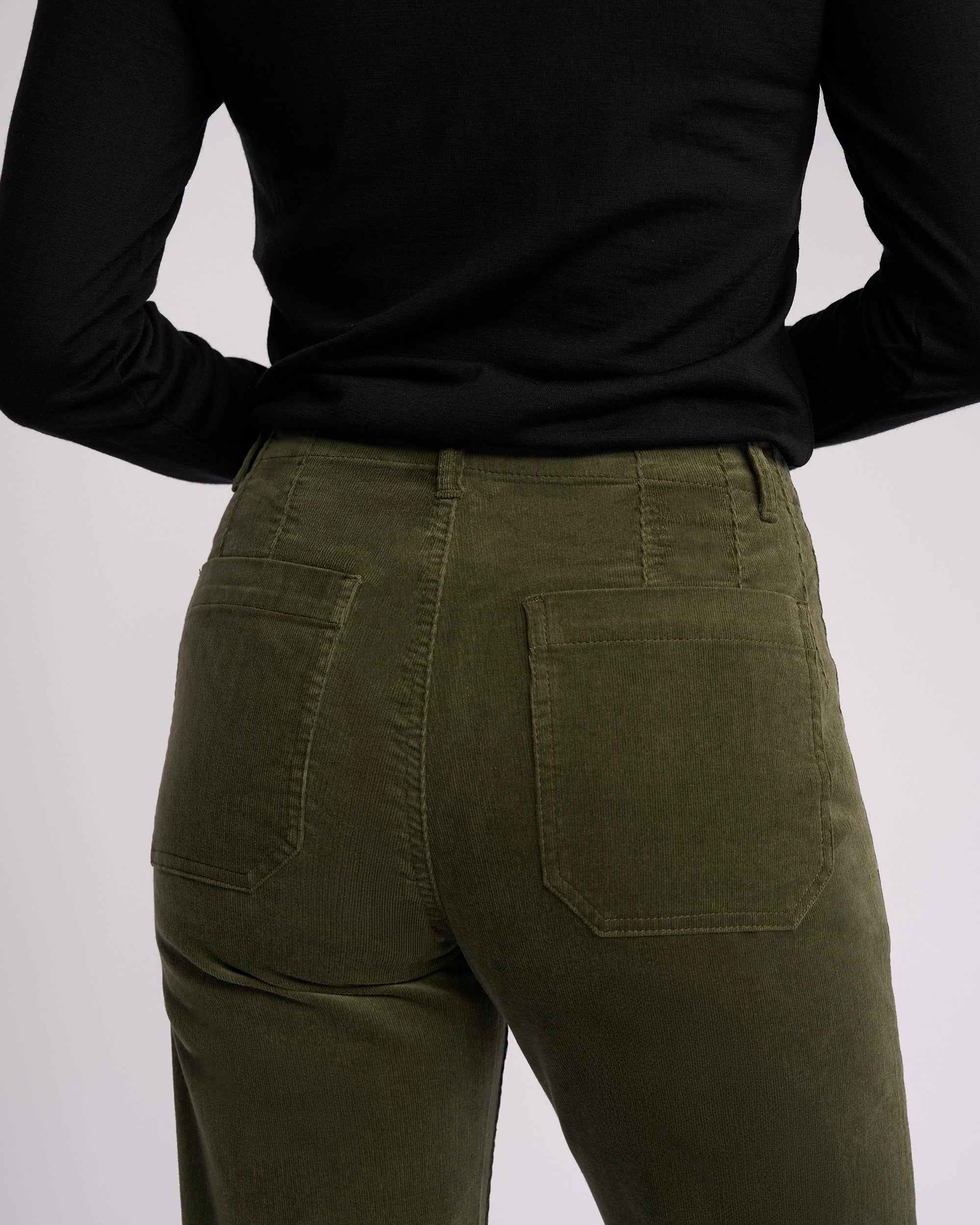 A person is seen from the back wearing Vassalli’s Daily Cord Pant in Deep Sage with a black long-sleeve top, hands on hips.
