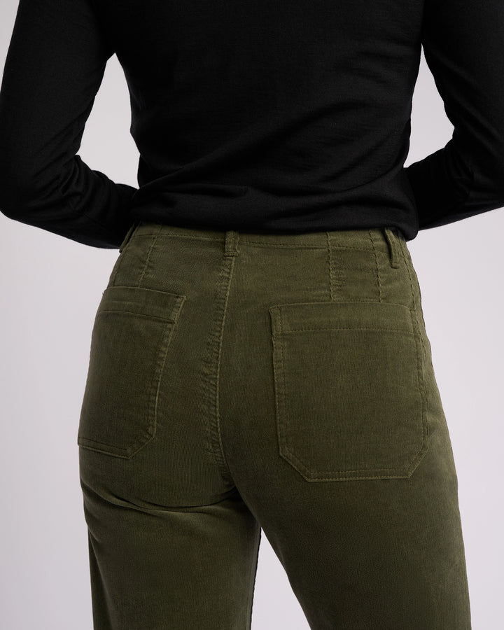 A person is seen from the back wearing Vassalli’s Daily Cord Pant in Deep Sage with a black long-sleeve top, hands on hips.