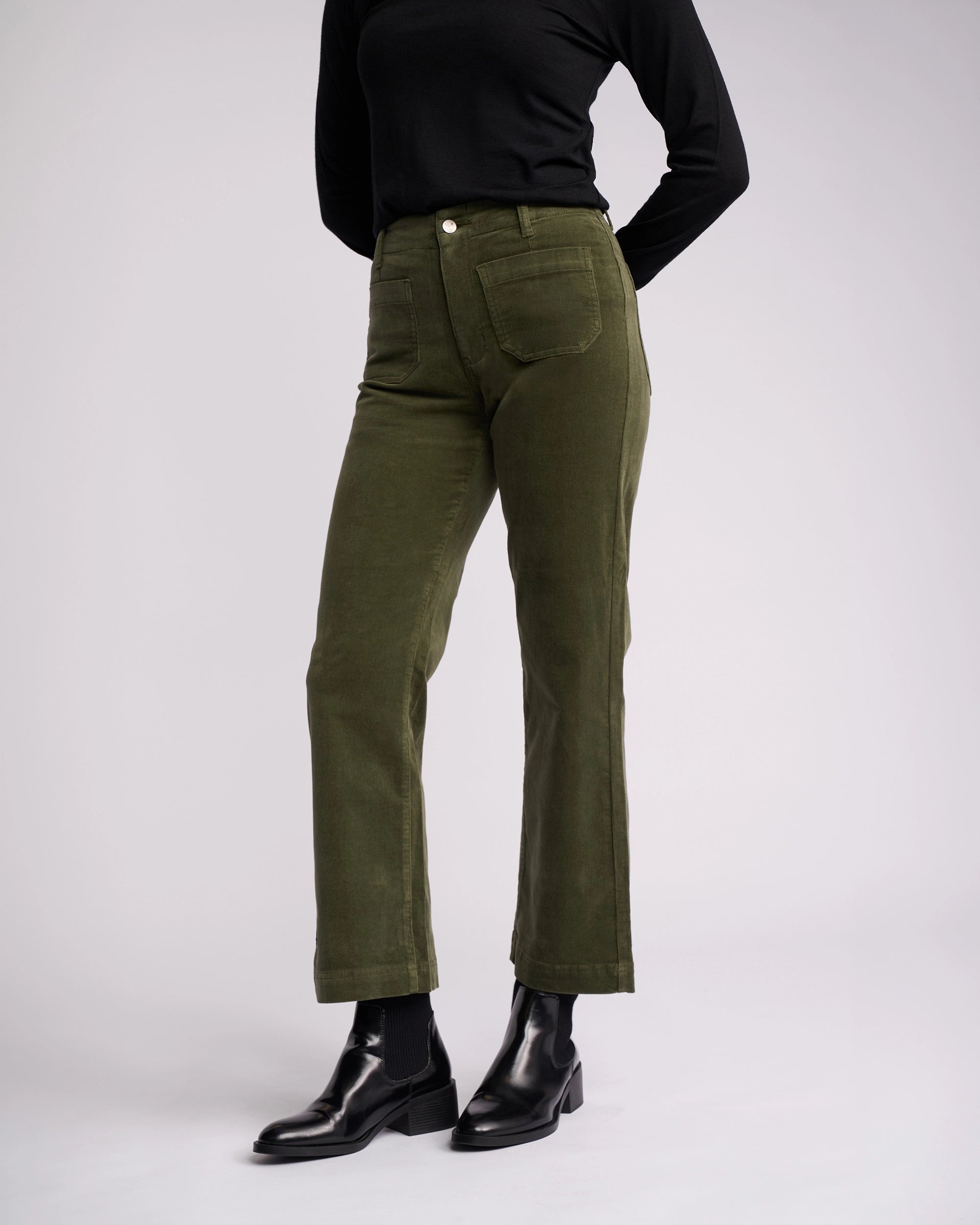 Model wearing Vassalli Daily Cord Pant in Deep Sage, paired with a black long-sleeve top, black socks, and black ankle boots, standing against a plain light background with hands behind their back.