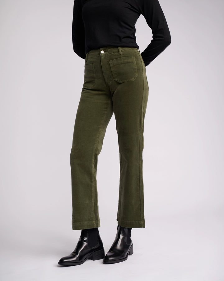 Model wearing Vassalli Daily Cord Pant in Deep Sage, paired with a black long-sleeve top, black socks, and black ankle boots, standing against a plain light background with hands behind their back.