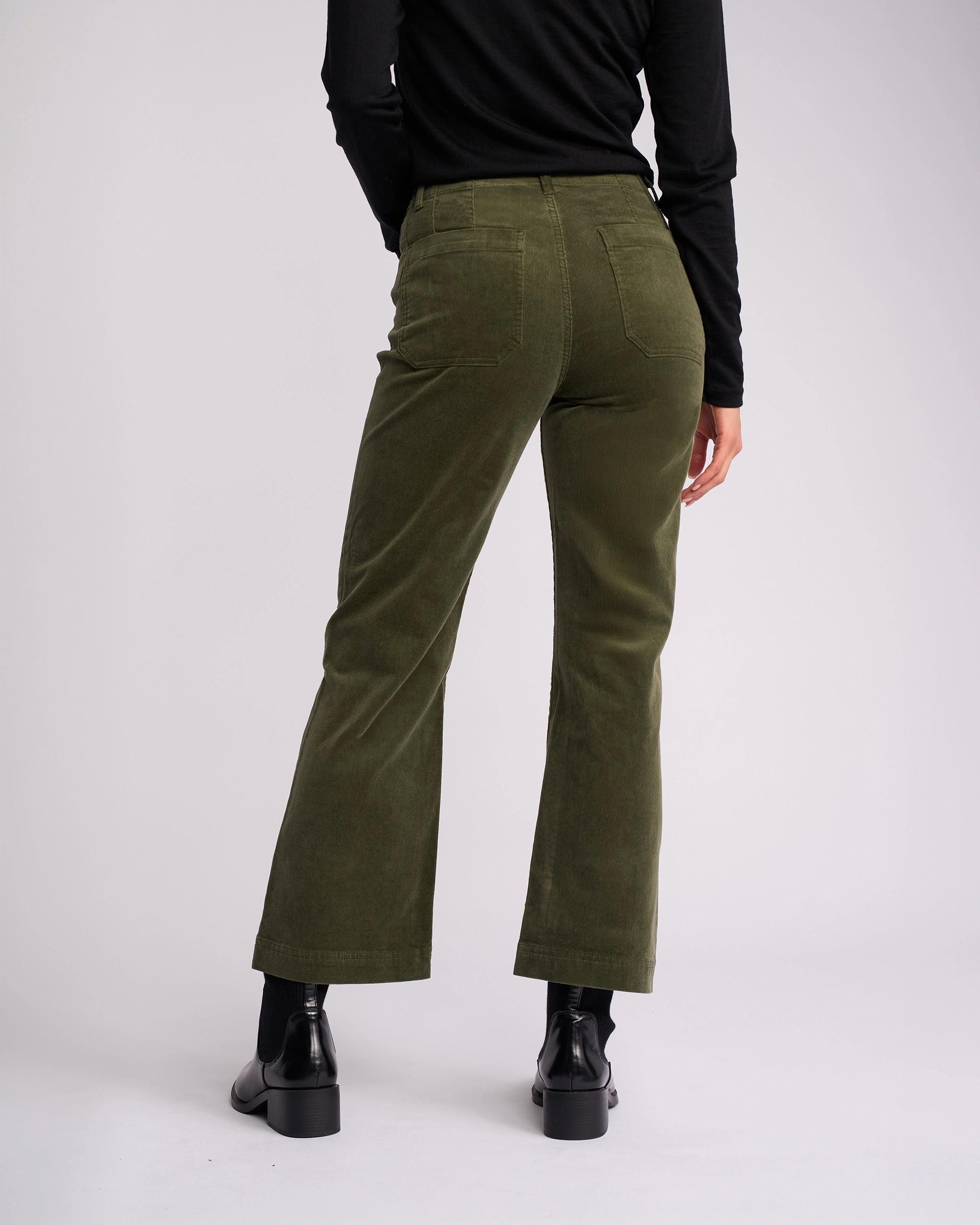 A person stands with their back to the camera, wearing Vassalli's Daily Cord Pant in Deep Sage, paired with a black long-sleeve top and black ankle boots, set against a plain light background.