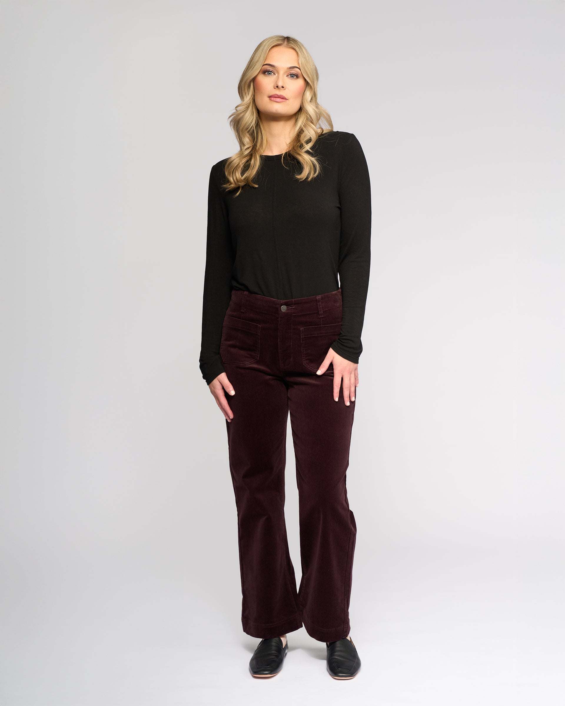 A woman with long blonde hair stands against a light background, wearing the Vassalli Daily Cord Pant in Merlot, a black long-sleeve top, and black shoes. She faces the camera with a neutral expression.