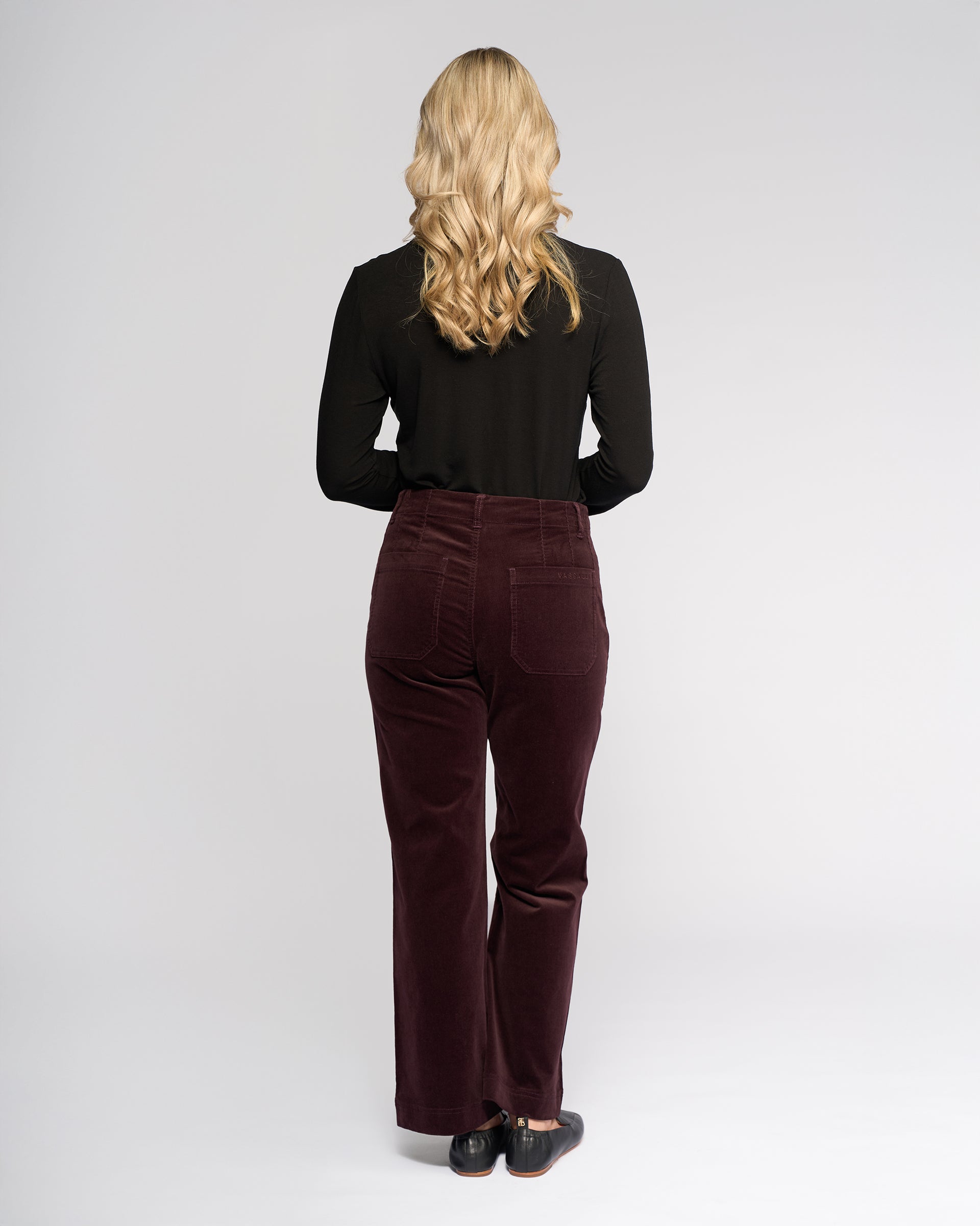 A woman with long, wavy blonde hair stands facing away, wearing a black top and Vassalli Daily Cord Pant in Merlot, paired with black shoes against a plain light gray background.