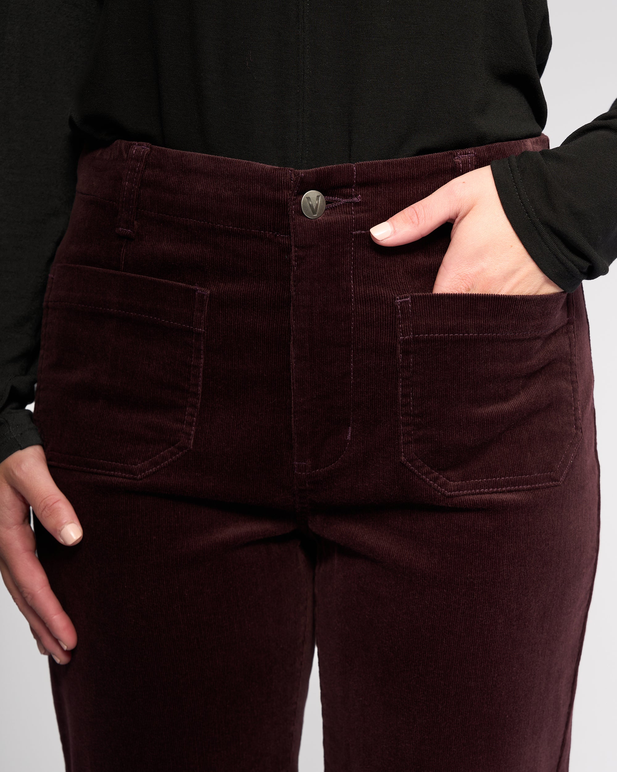 A person wearing Vassalli's Daily Cord Pant in Merlot, paired with a black top. One hand is in their pocket, the other by their side. The image focuses on the lower torso and upper legs.