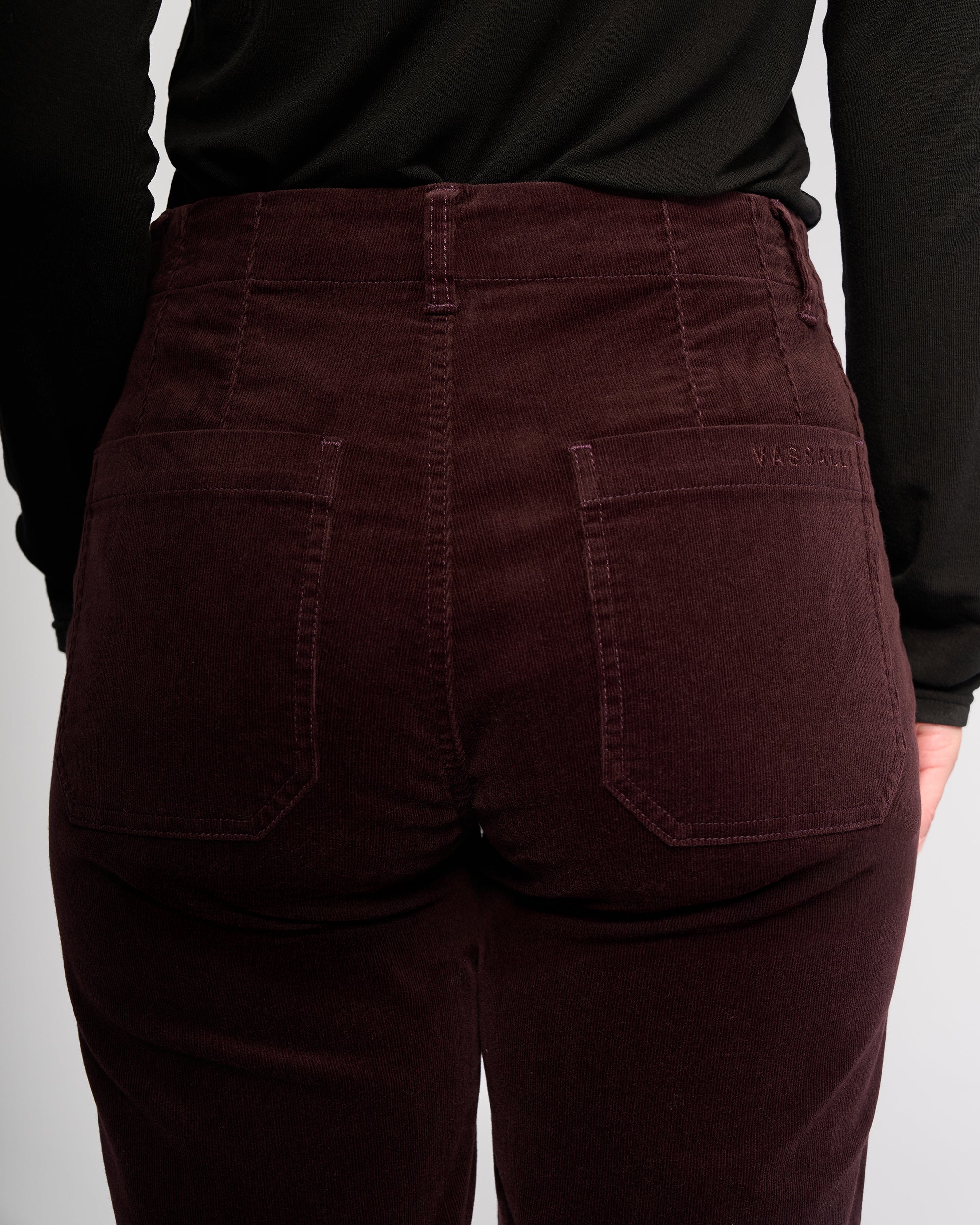 A person shown from the back waist down wearing Vassalli's Daily Cord Pant in Merlot, a high-waisted corduroy ankle-grazer with two back pockets, visible stitching, and a black top.