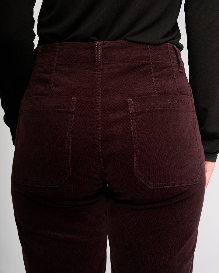 A person shown from the back waist down wearing Vassalli's Daily Cord Pant in Merlot, a high-waisted corduroy ankle-grazer with two back pockets, visible stitching, and a black top.