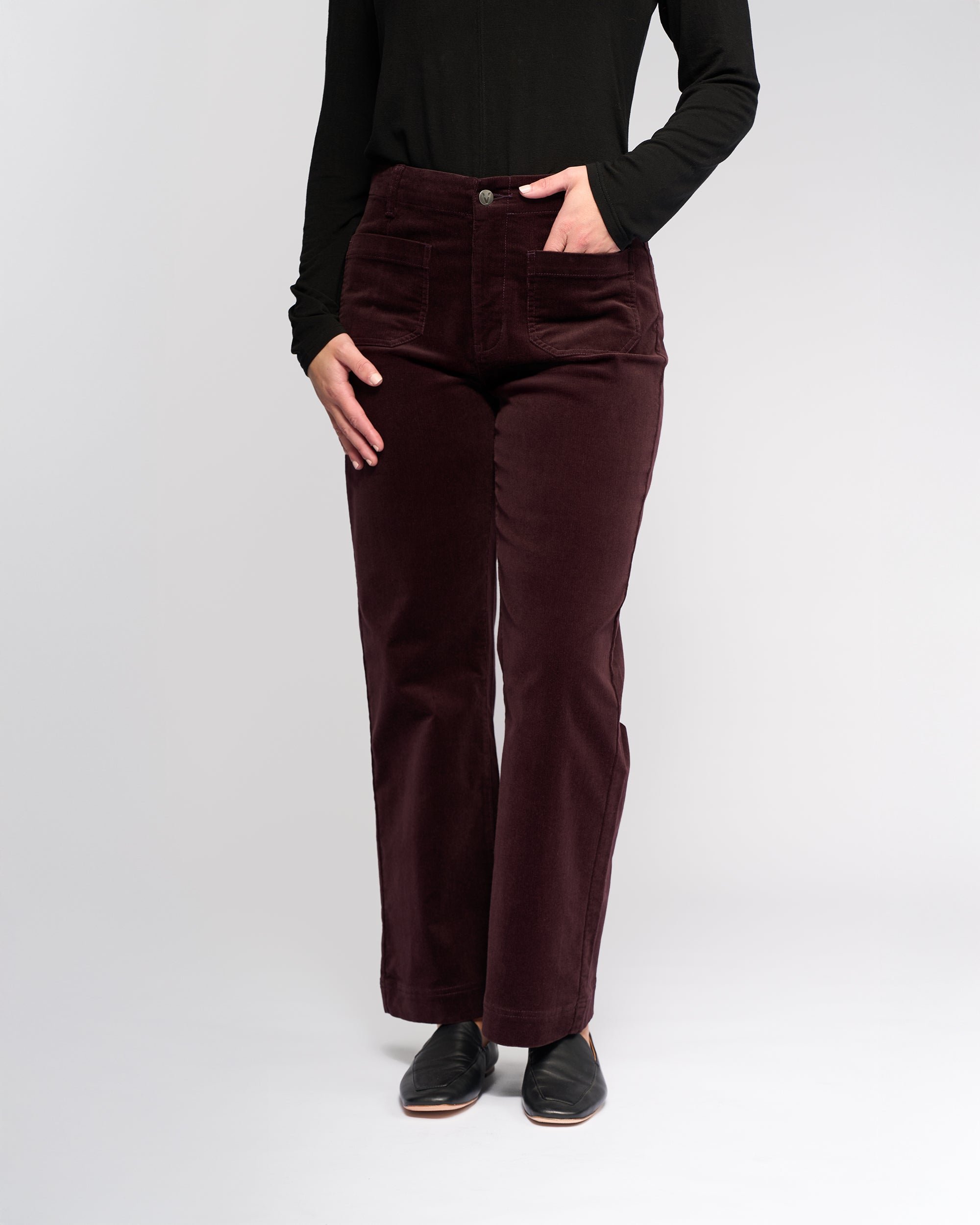 A person wearing a black long-sleeve top, black shoes, and Vassalli Daily Cord Pant in Merlot stands against a plain light background. The person's face is not visible.