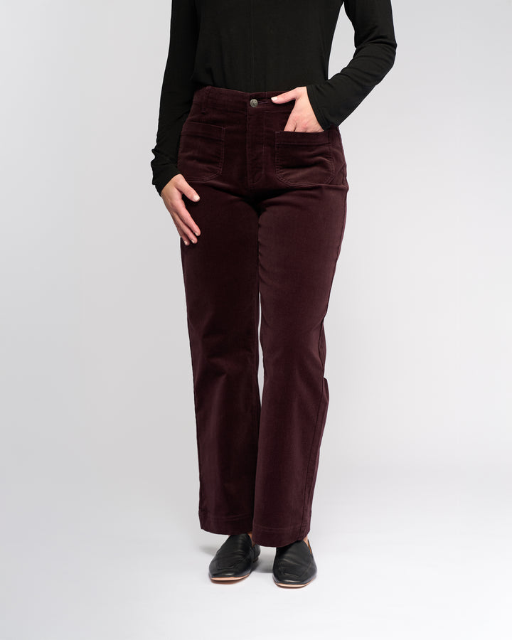 A person wearing a black long-sleeve top, black shoes, and Vassalli Daily Cord Pant in Merlot stands against a plain light background. The person's face is not visible.