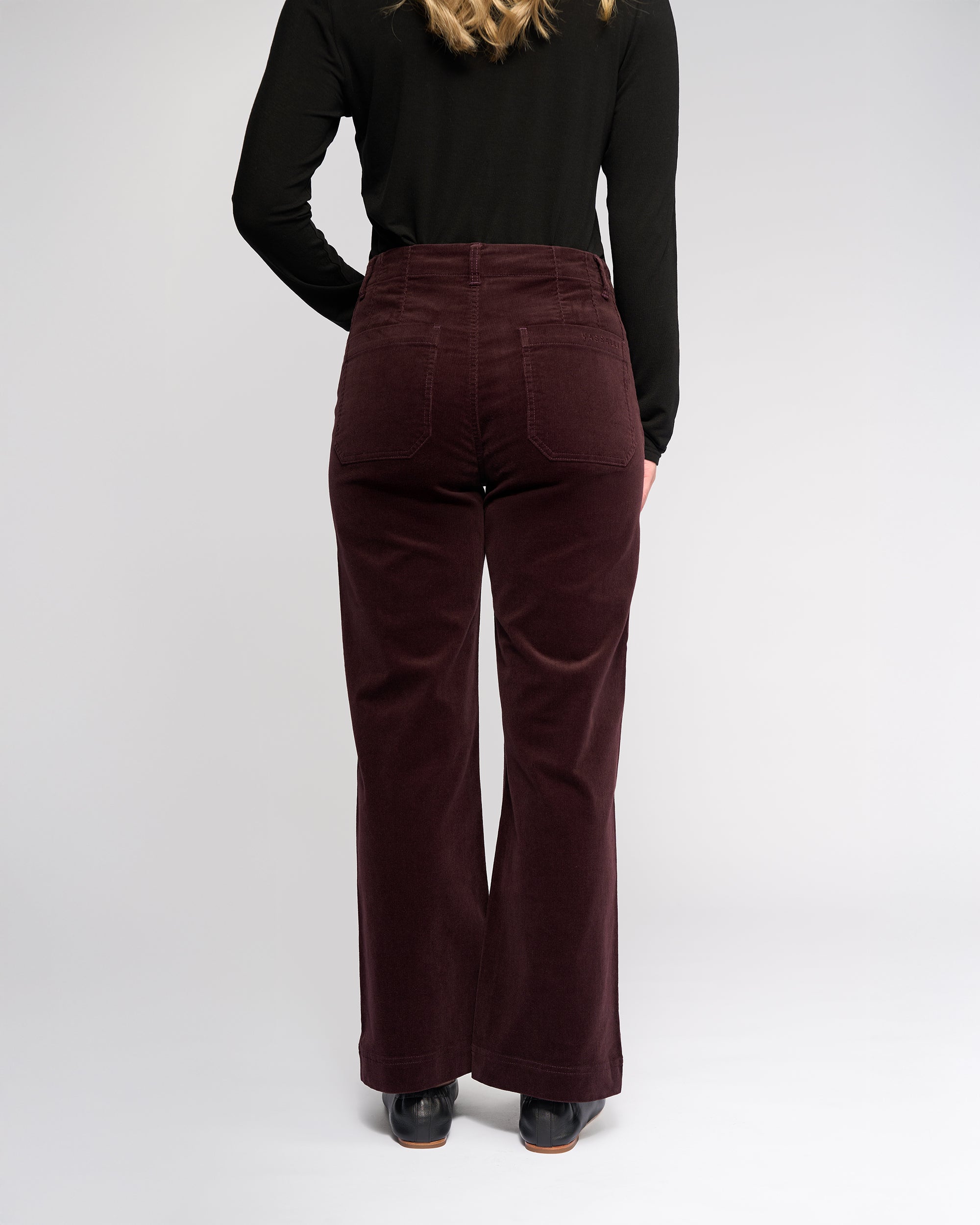 A person stands facing away in a black long-sleeve top and Vassalli Daily Cord Pant - Merlot, featuring pockets. The plain, light gray background highlights the corduroy ankle-grazer style.
