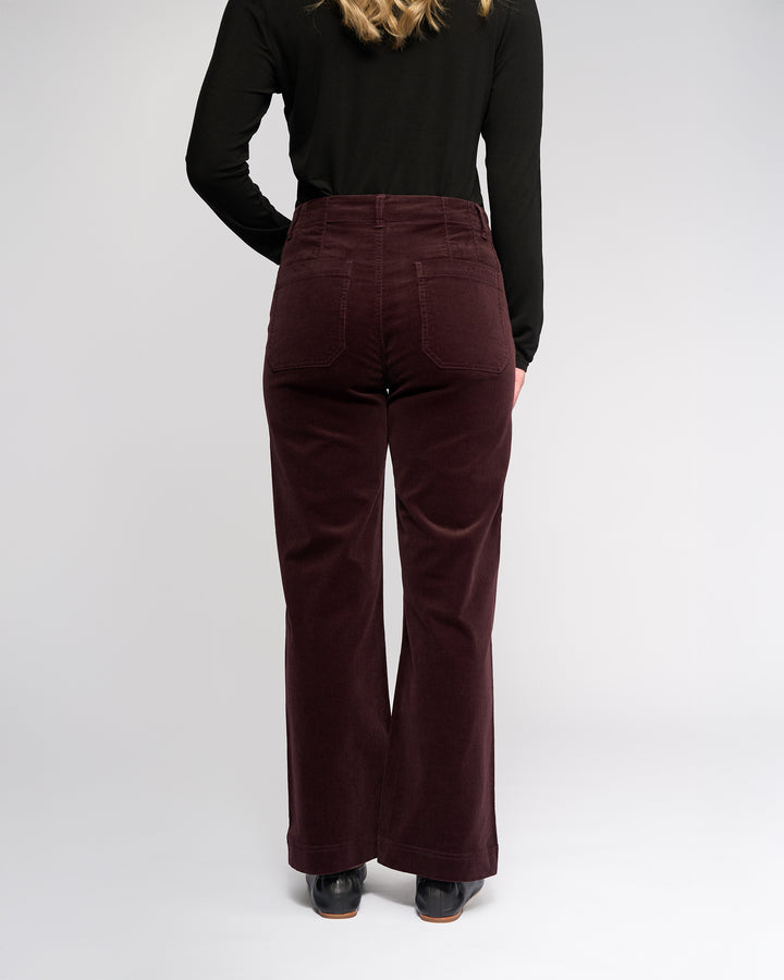 A person stands facing away in a black long-sleeve top and Vassalli Daily Cord Pant - Merlot, featuring pockets. The plain, light gray background highlights the corduroy ankle-grazer style.