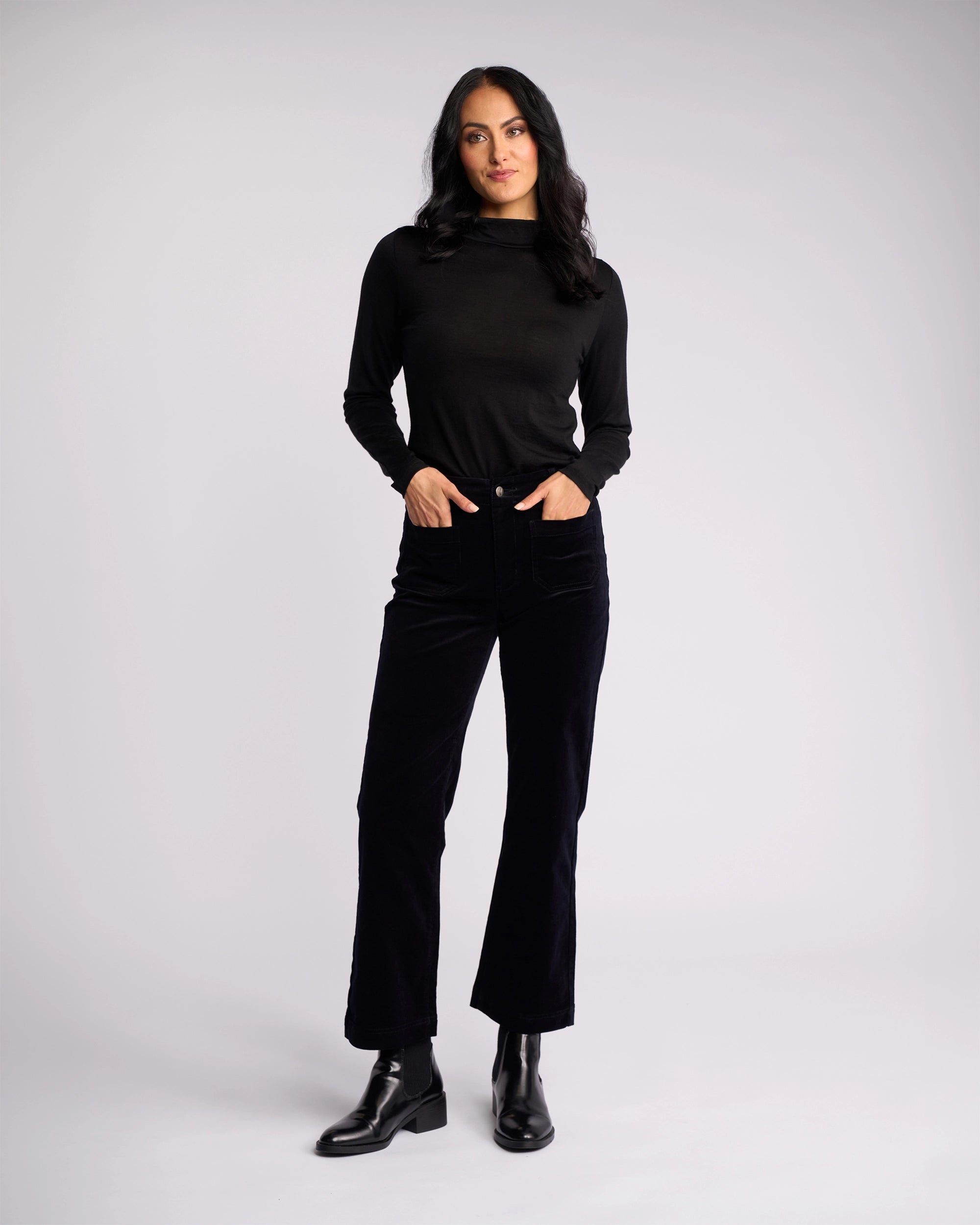 A woman with long dark hair wears the Vassalli Daily Cord Pant in Midnight, a black long-sleeve top, and black ankle boots as she stands with hands in her pockets against a plain light background.