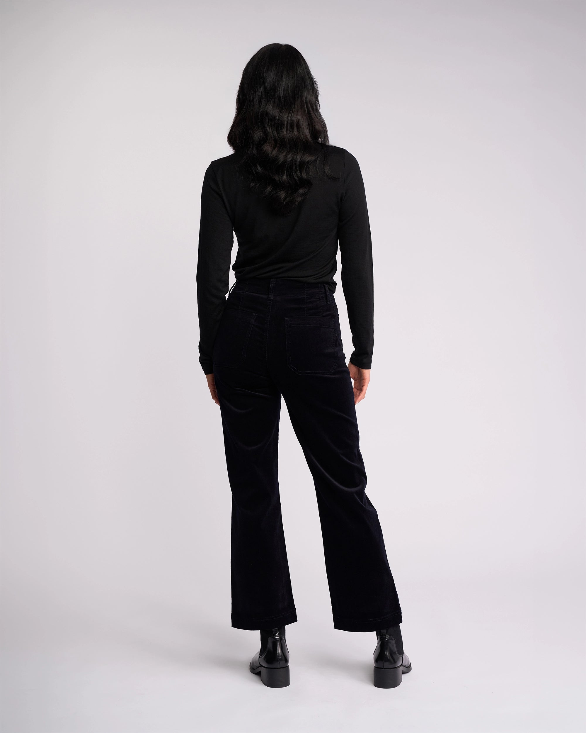 A person with long dark wavy hair, wearing a black long-sleeve top and Vassalli Daily Cord Pant - Midnight, paired with black ankle boots, stands facing away from the camera against a plain light background.