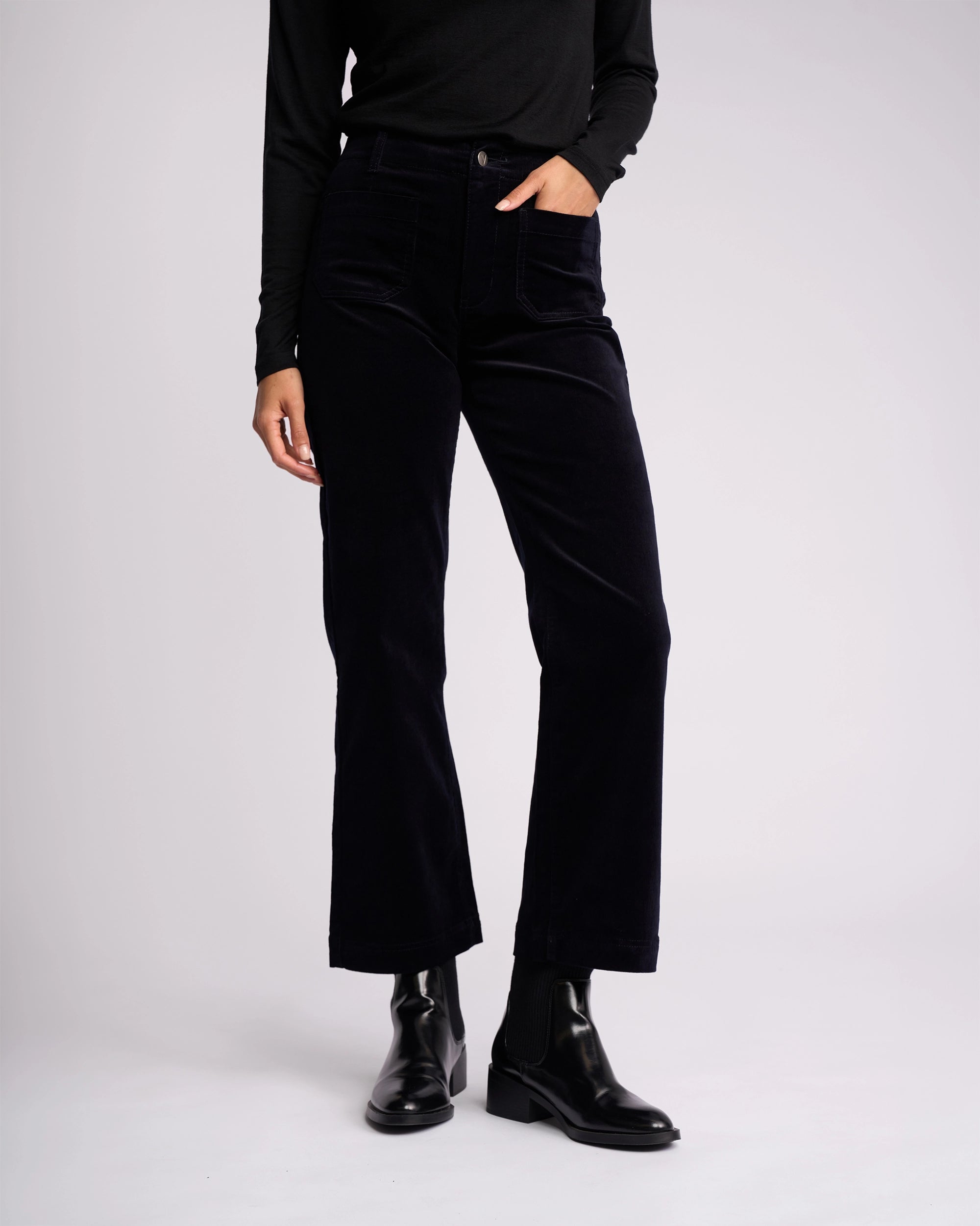 A person wears Vassalli’s Daily Cord Pant - Midnight, a long-sleeve black top, and black ankle boots, standing against a plain light background with one hand in their pocket. Their face is not shown.