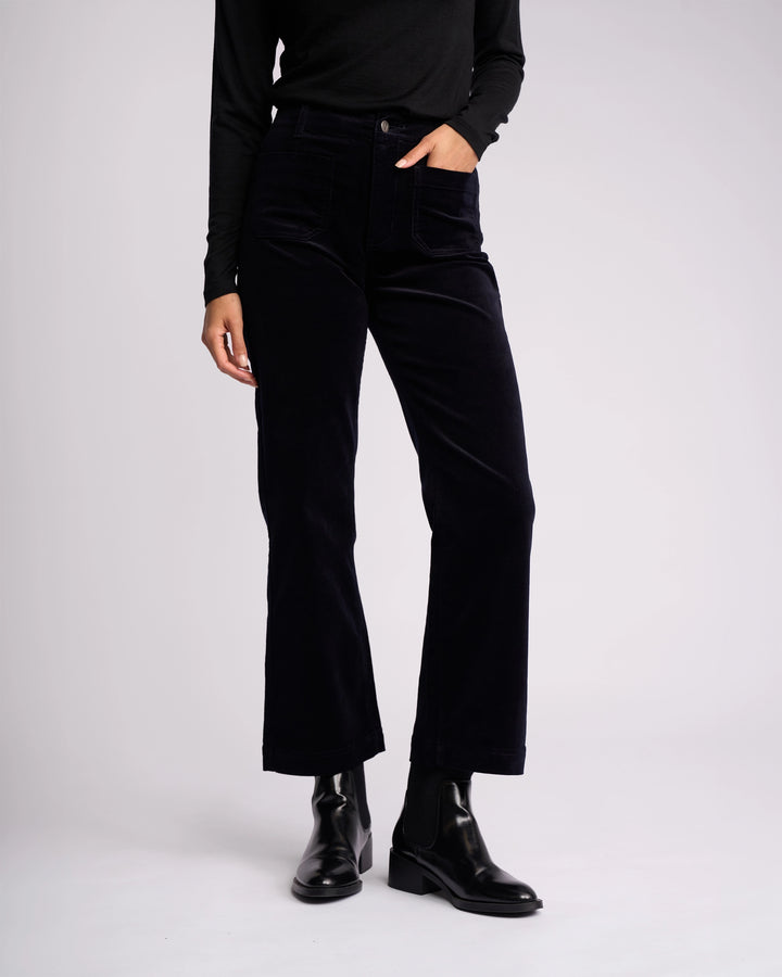 A person wears Vassalli’s Daily Cord Pant - Midnight, a long-sleeve black top, and black ankle boots, standing against a plain light background with one hand in their pocket. Their face is not shown.