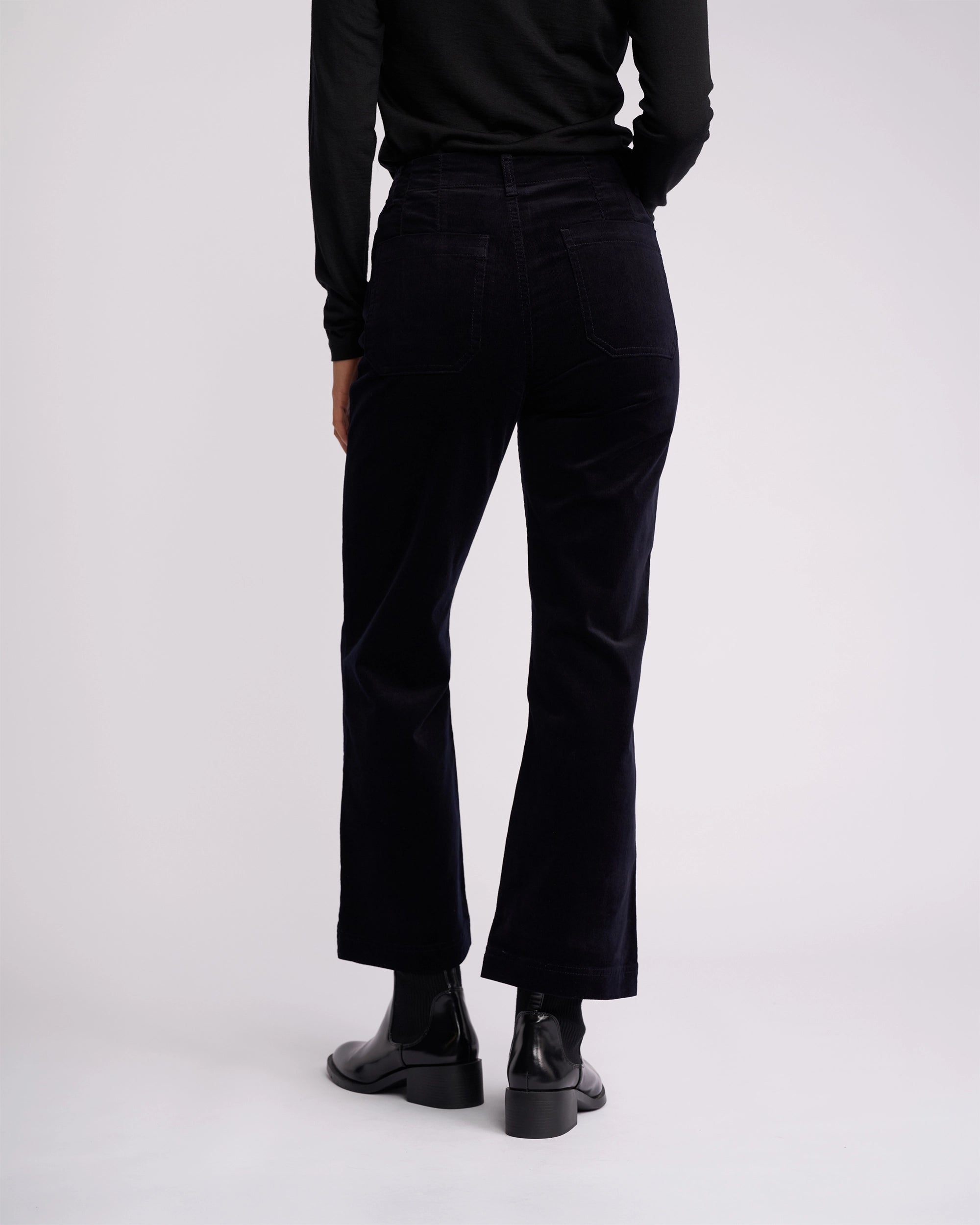 A person is shown from the back against a white background, wearing a black long-sleeve top, black ankle boots, and Vassalli’s Daily Cord Pant in Midnight—a straight-leg, high-waisted style with back pockets.