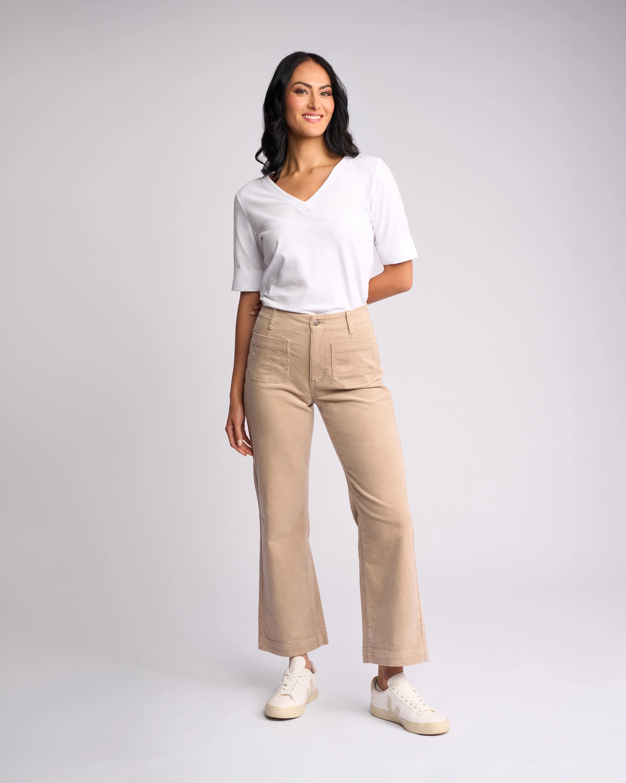 A woman stands smiling against a plain light background, wearing a white V-neck shirt, Vassalli's Daily Cord Pant - Oat, and white sneakers. Her hair is loose and wavy.