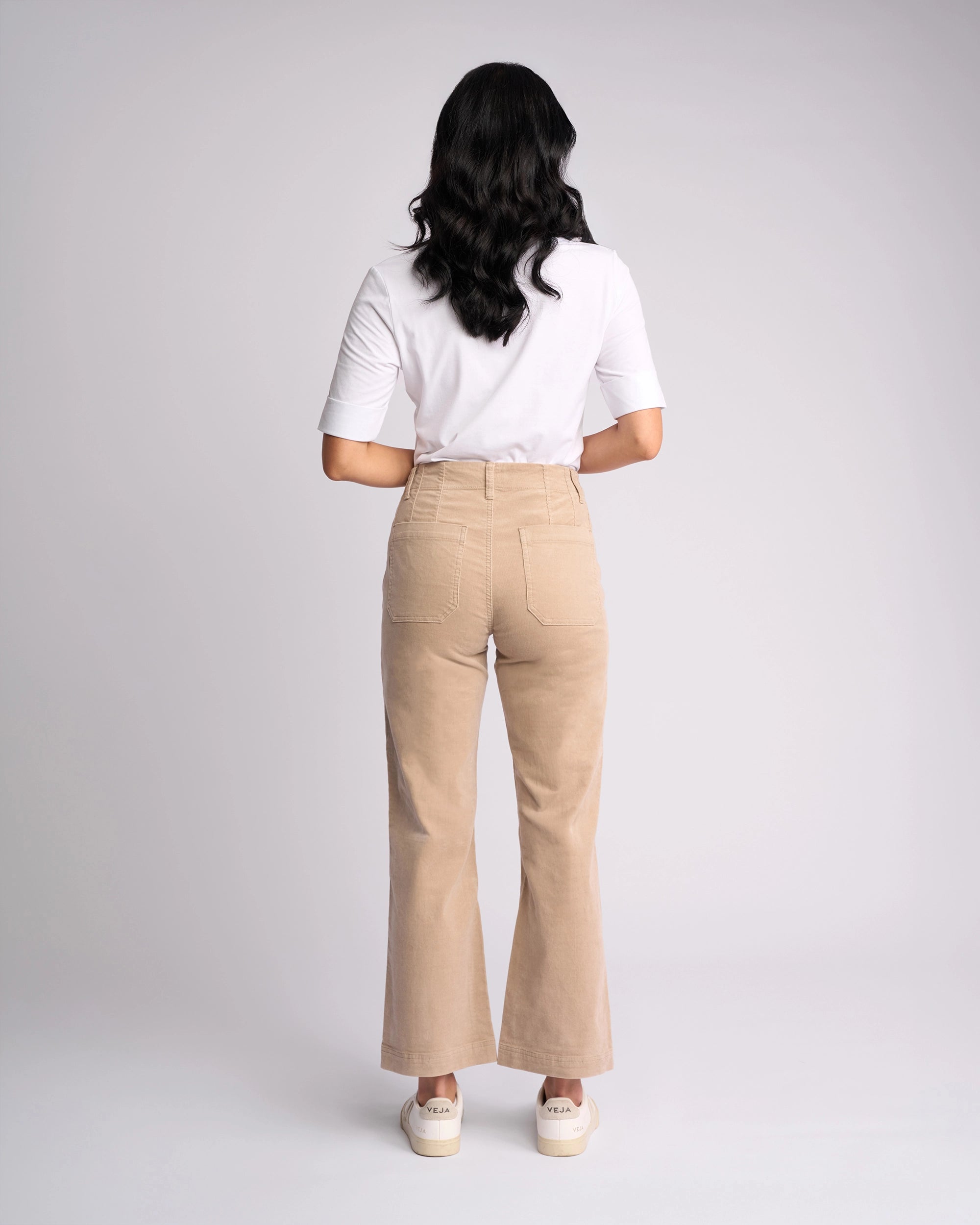 A person with long dark hair faces away, wearing a white short-sleeve shirt, Vassalli Daily Cord Pant - Oat (high-waisted, straight-leg), and white sneakers against a plain light gray background.
