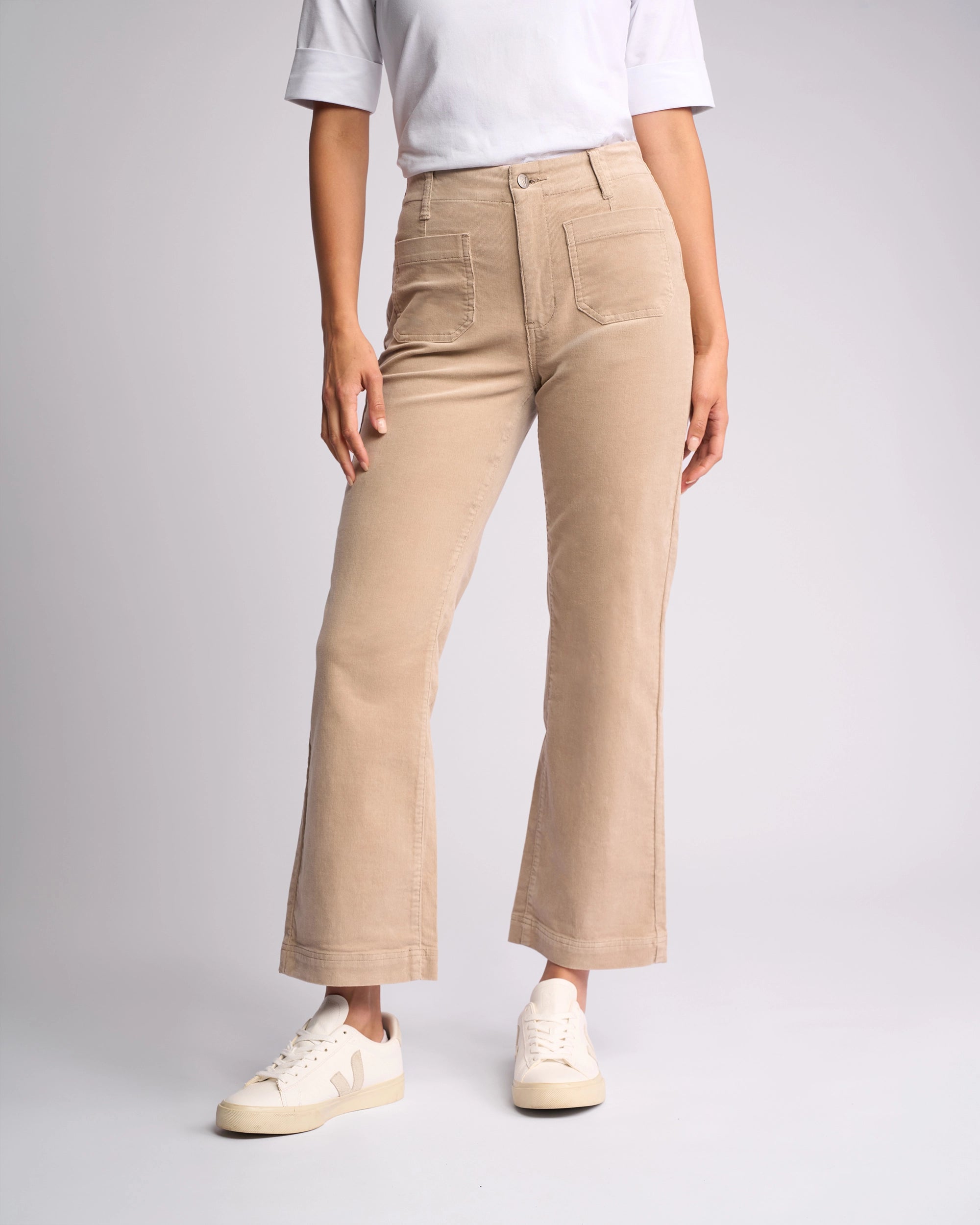 A person wearing Vassalli's Daily Cord Pant in oat, featuring a high waist, straight leg, and front pockets, paired with a white short-sleeve top and white sneakers, stands against a plain light background.