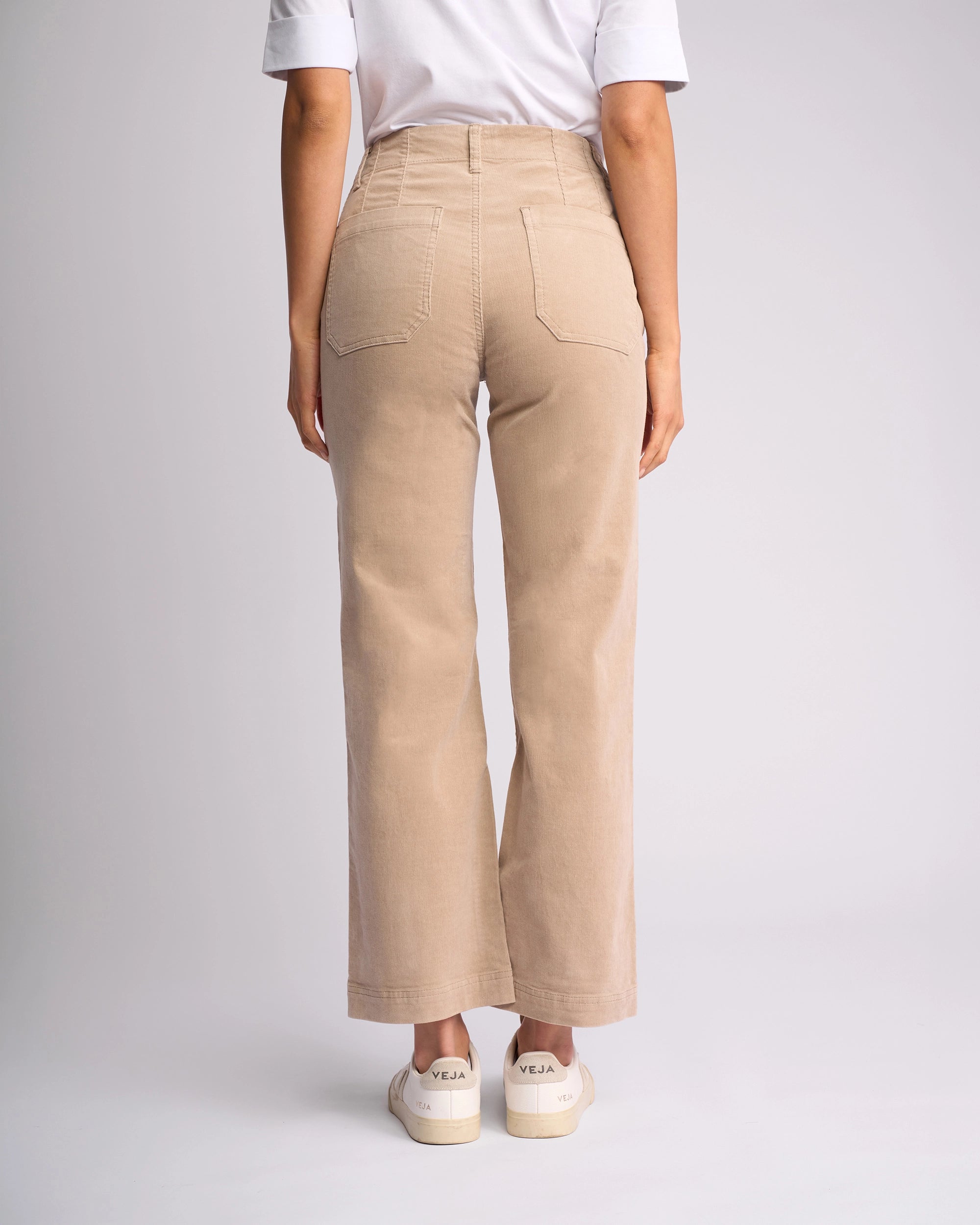A person stands facing away in Vassalli’s Daily Cord Pant - Oat, styled with a white t-shirt and white sneakers featuring VEJA branding. The background is plain and light-colored.