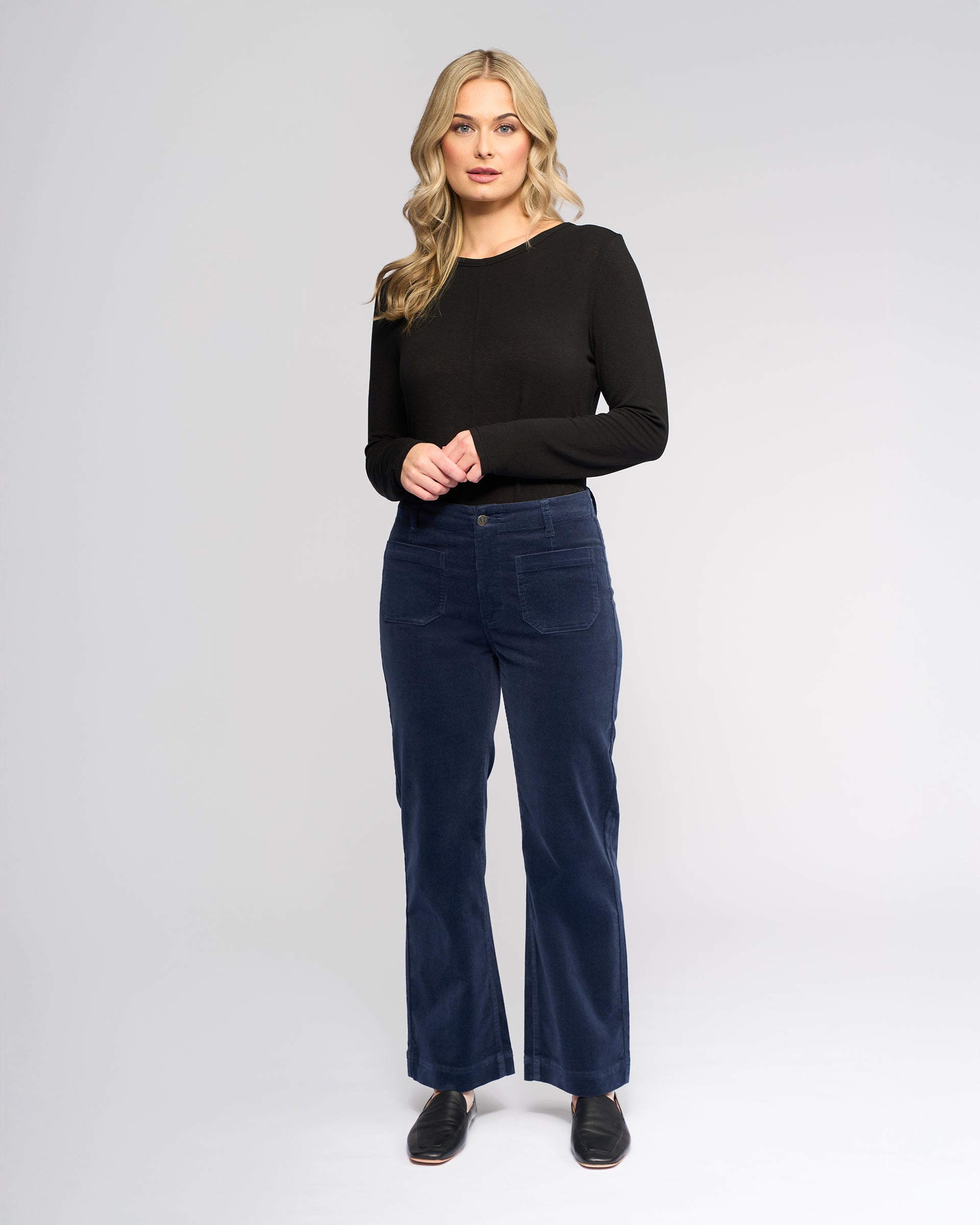 A woman with long blonde hair wears a black long-sleeve top, Vassalli Daily Cord Pant in Slate, and black shoes, standing against a plain light gray background.