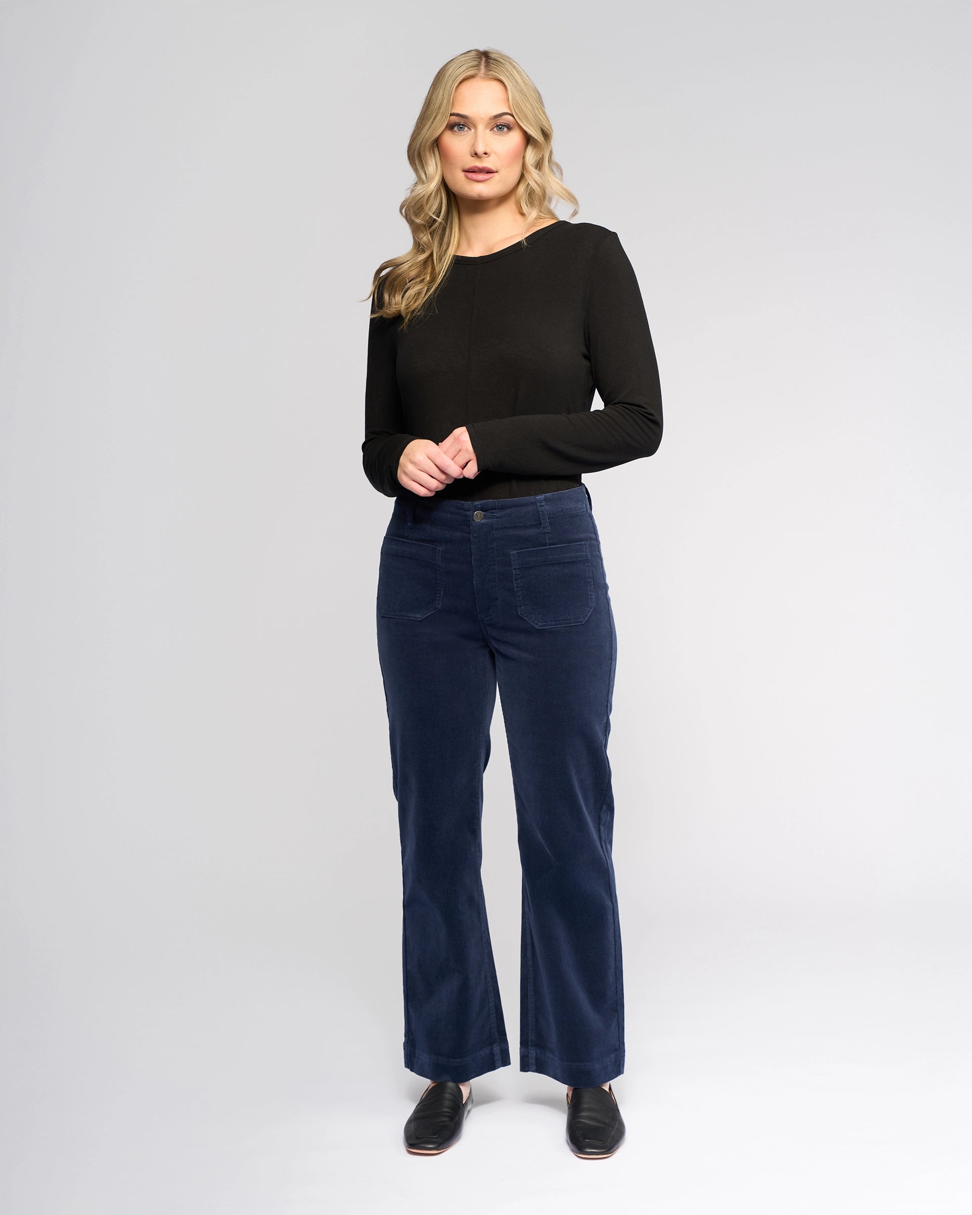 A woman with long blonde hair, wearing a black long-sleeve top and Vassalli Daily Cord Pant in Slate, stands facing the camera against a plain light gray background.