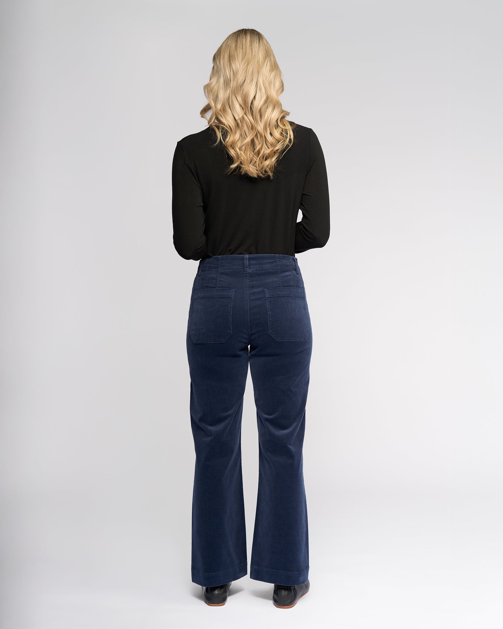A person with long, wavy blonde hair stands facing away, wearing a black long-sleeve top and Vassalli's Daily Cord Pant in Slate, which are blue high-waisted flared corduroy pants, along with black shoes against a plain light background.