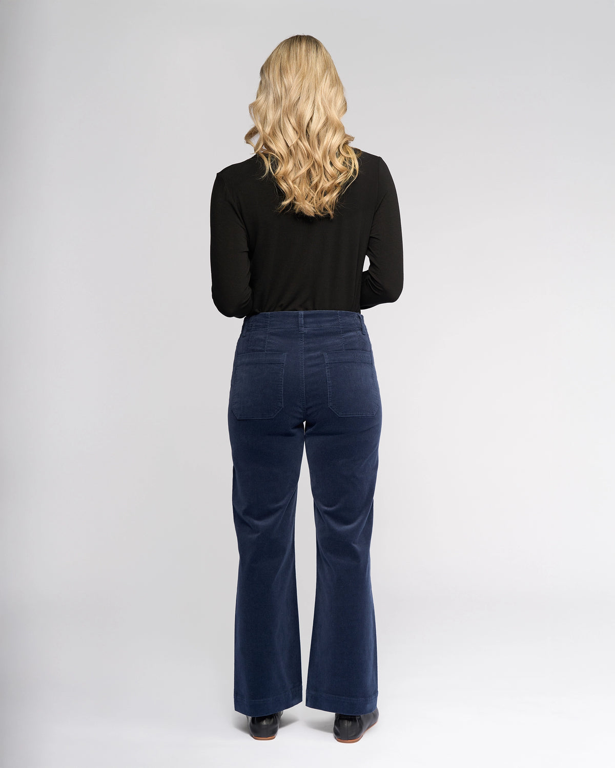 A person with long blonde hair stands facing away, wearing a black long-sleeve top, Vassalli Daily Cord Pant - Slate, and black shoes against a plain light gray background.