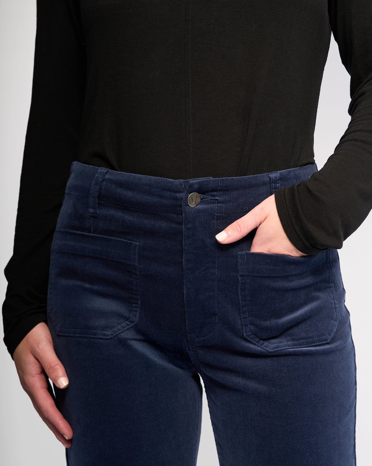 Wearing a long-sleeve black top and Vassalli's Daily Cord Pant in Slate, featuring a high waist, straight leg, and front pockets. One hand is in a pocket while the other rests by their side.