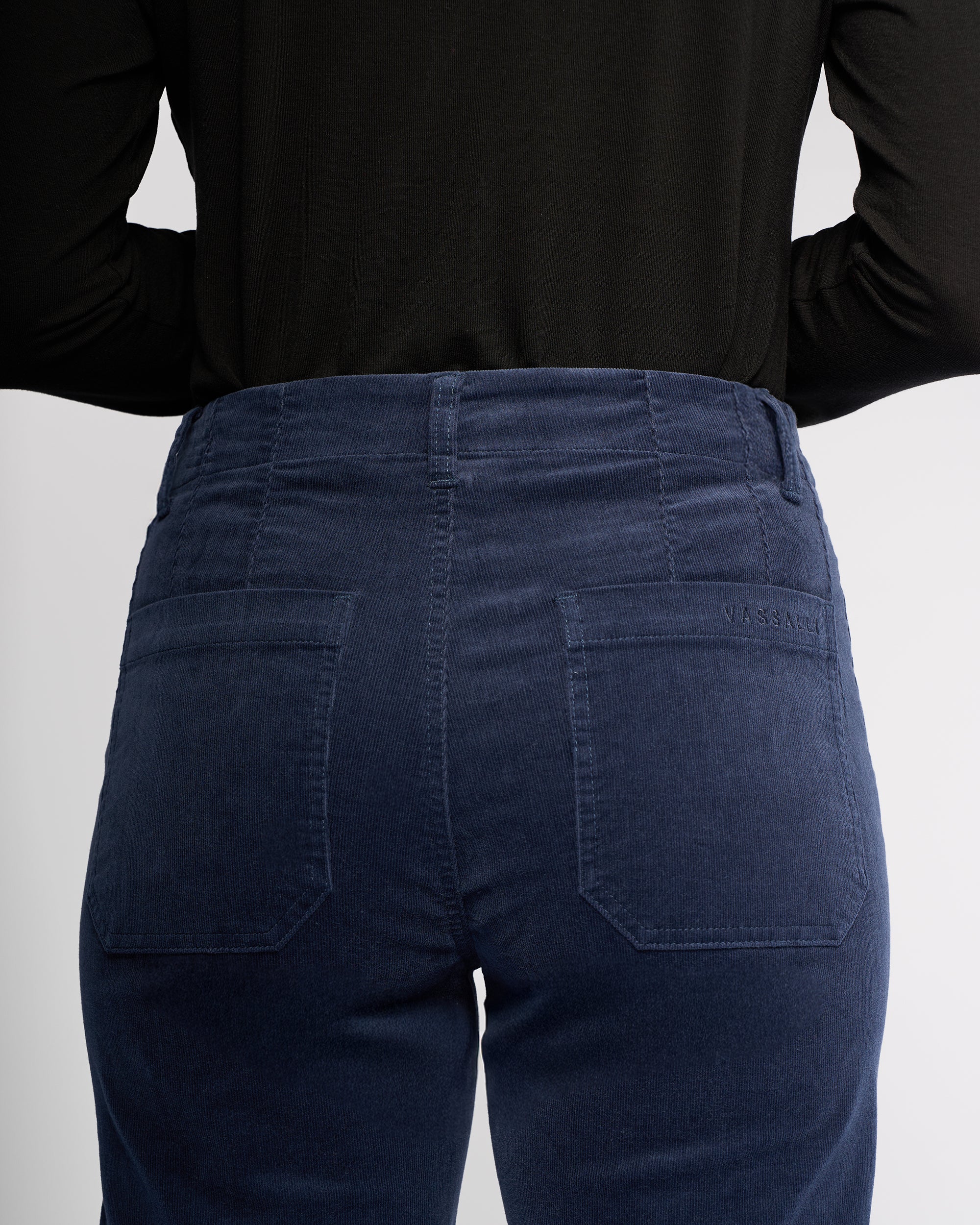 A person, shown from the back, highlights the waistband and back pockets of the Vassalli Daily Cord Pant - Slate, a high-waisted blue straight-leg ankle-grazer style.