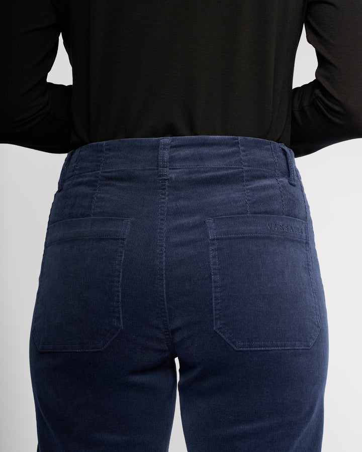 A person, shown from the back, highlights the waistband and back pockets of the Vassalli Daily Cord Pant - Slate, a high-waisted blue straight-leg ankle-grazer style.