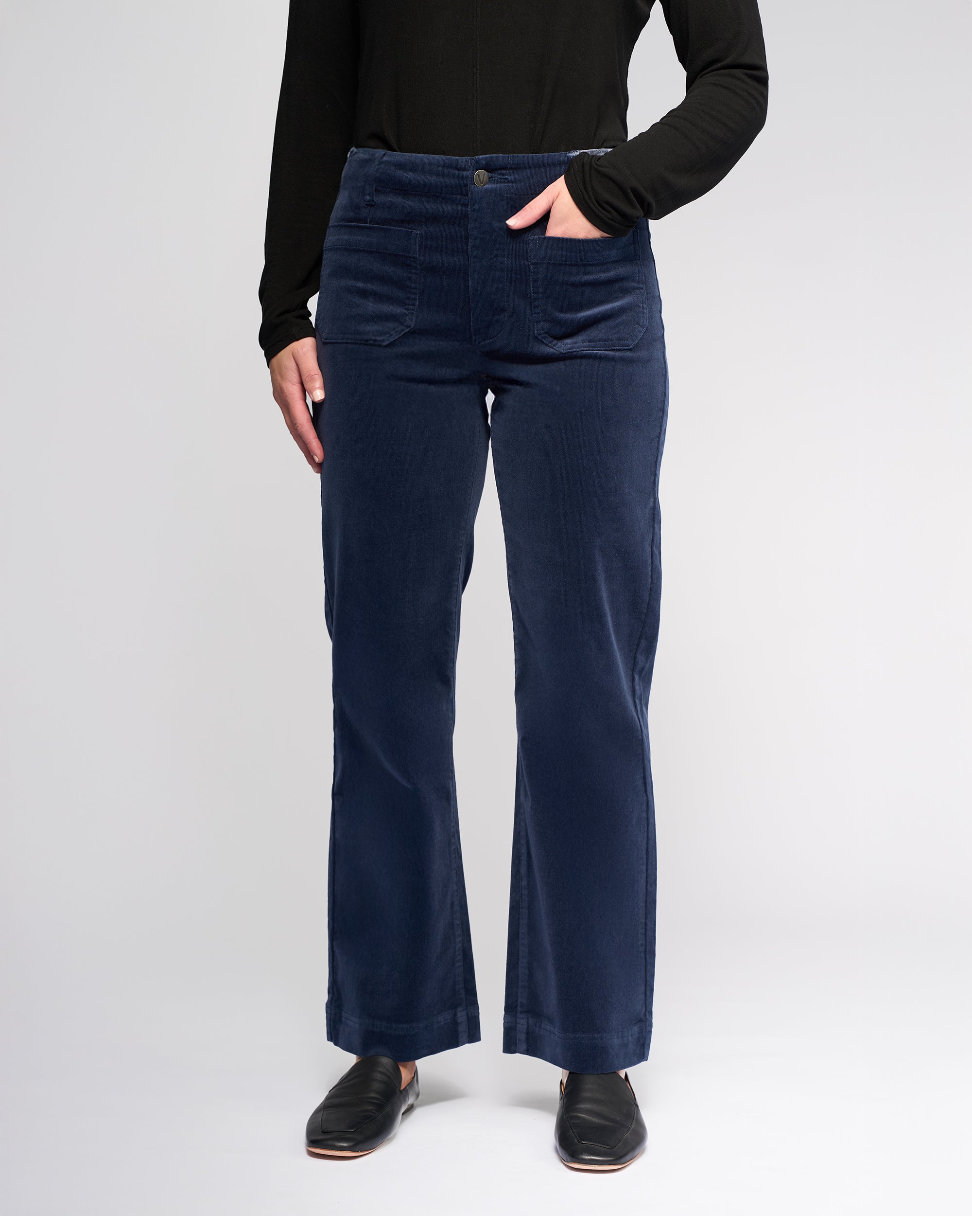 A person wears the Vassalli Daily Cord Pant in Slate with one hand in their pocket, paired with a black long-sleeve top, standing against a plain light background. Only their body from shoulders down is shown.