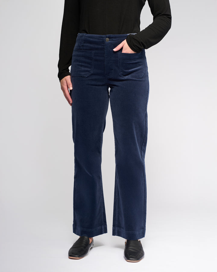 A person wears the Vassalli Daily Cord Pant in Slate with one hand in their pocket, paired with a black long-sleeve top, standing against a plain light background. Only their body from shoulders down is shown.