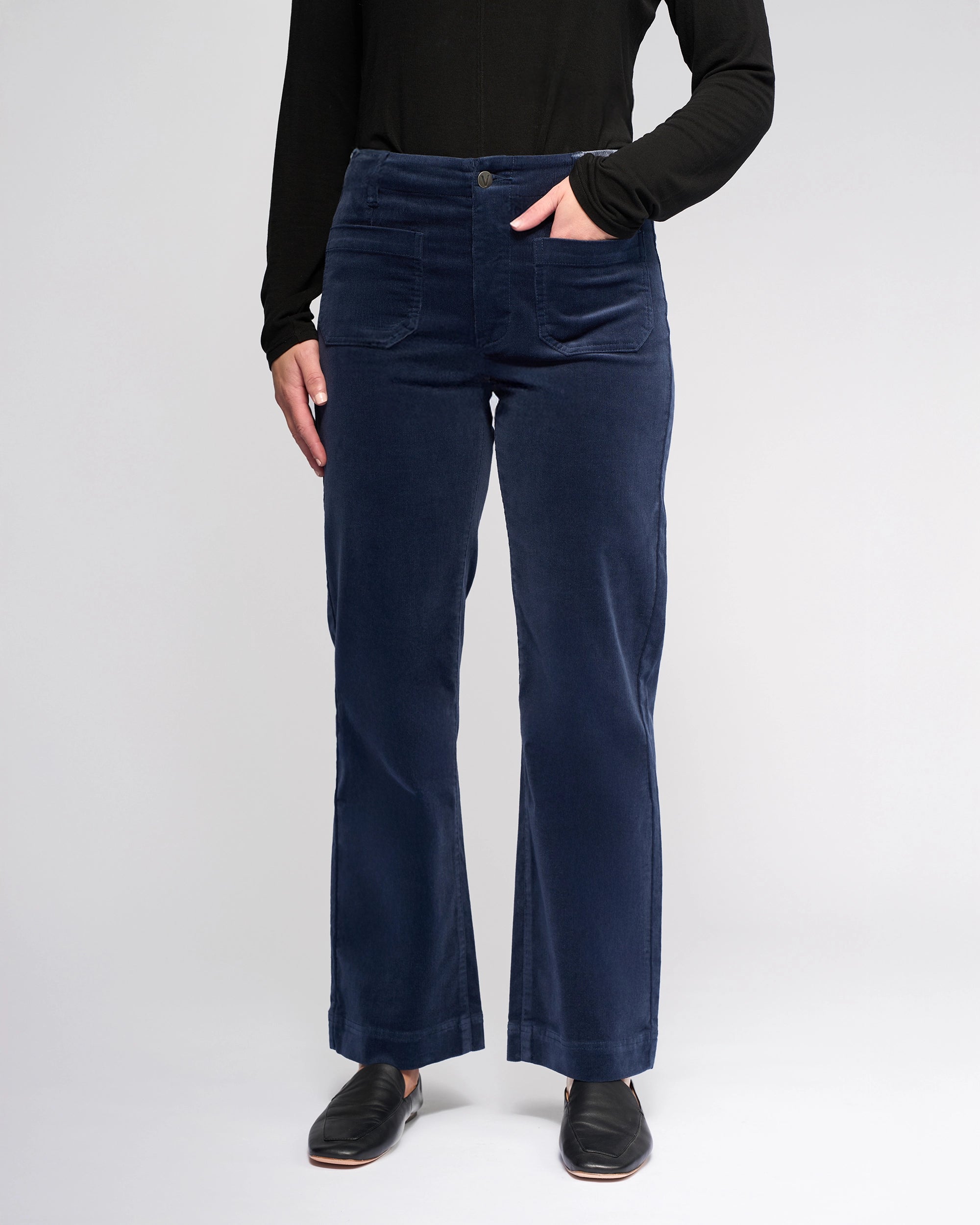 A person stands against a light background wearing a black long-sleeve top, Vassalli Daily Cord Pant in Slate, and black slip-on shoes, with one hand in a pocket.