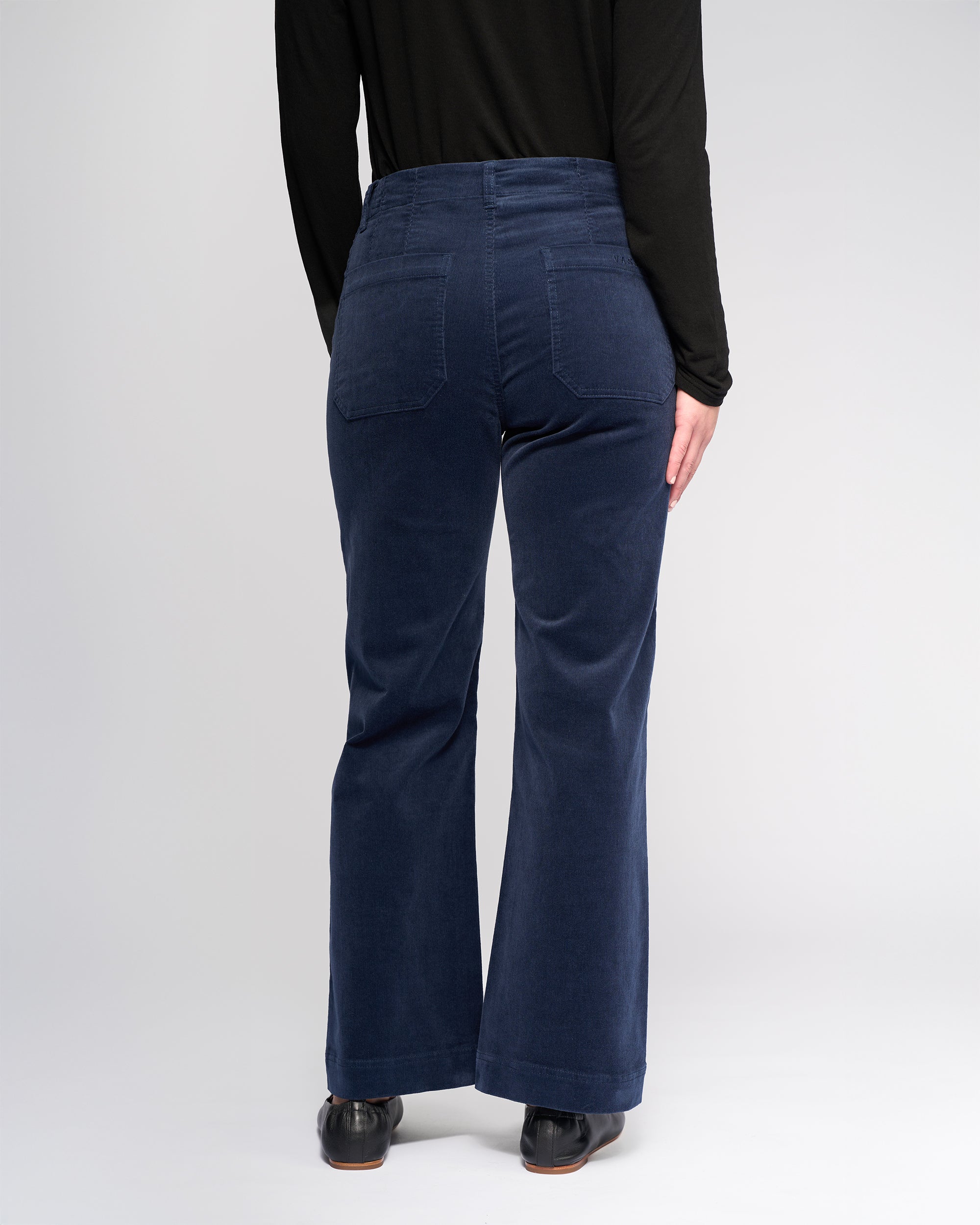 A person wearing Vassalli's Daily Cord Pant in Slate—a navy blue, wide-leg, straight-leg, ankle-grazer style with two back pockets—and a black long-sleeve top stands facing away from the camera.
