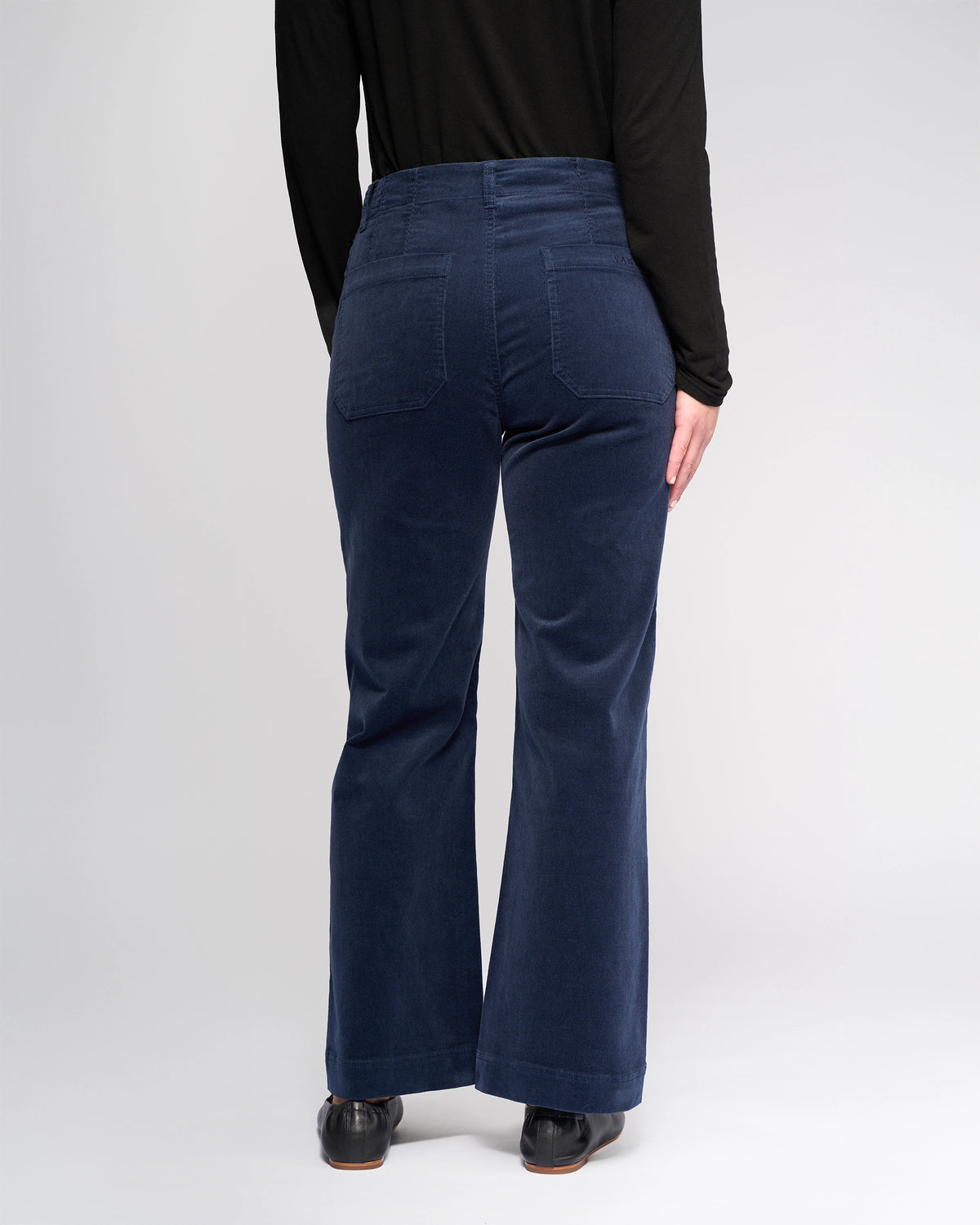 A person in Vassalli's Daily Cord Pant - Slate and a black long-sleeve top stands facing away from the camera against a plain, light background.