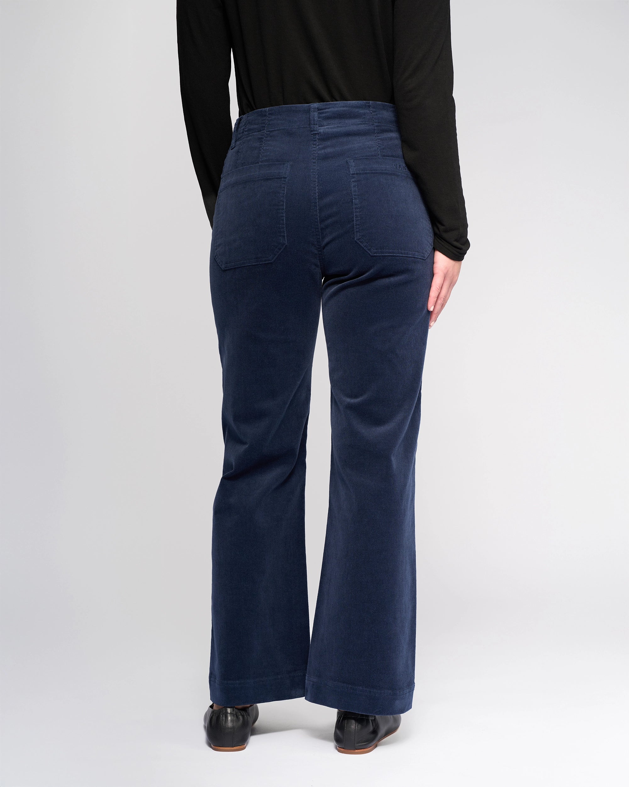 A person in Vassalli's Daily Cord Pant - Slate and a black long-sleeve top stands facing away from the camera against a plain, light background.