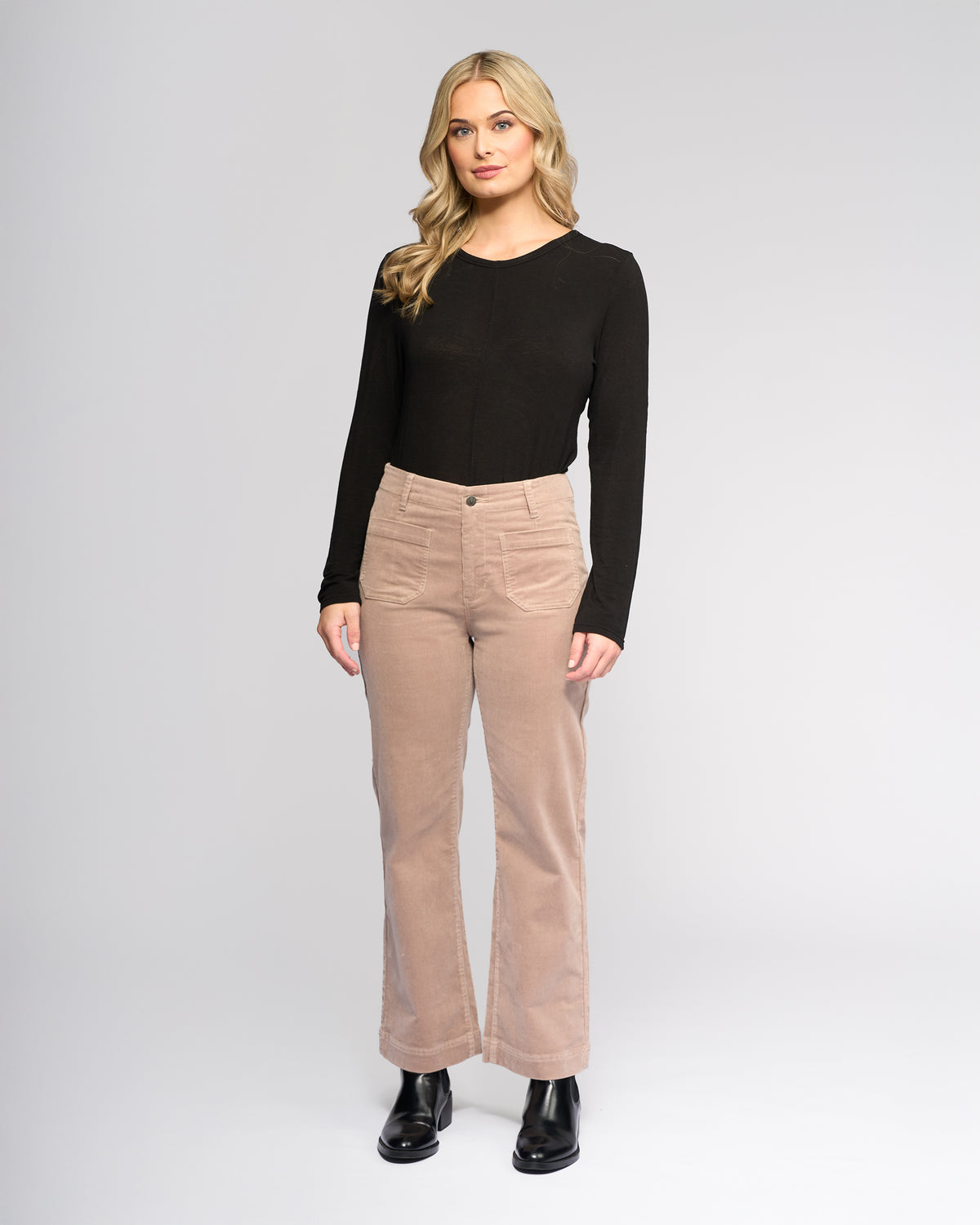 A woman with long blonde hair models the Vassalli Daily Cord Pant - Twig, styled with a black long-sleeve top and black boots, as she stands facing the camera against a plain light gray background.