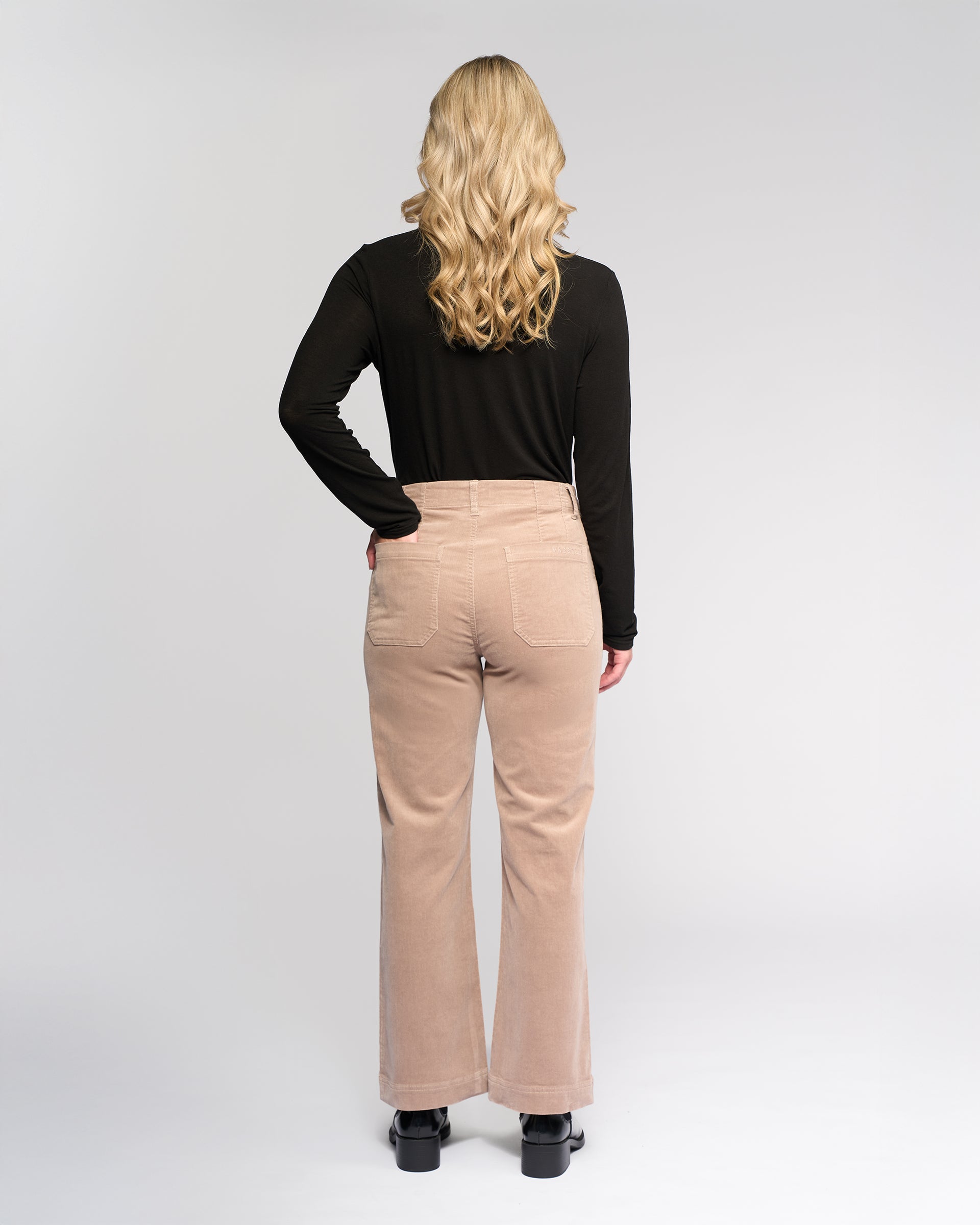 A woman with long, wavy blonde hair stands facing away against a plain light background, wearing a black top, black shoes, and Vassalli's Daily Cord Pant - Twig, beige high-waisted corduroy pants with patch pockets.