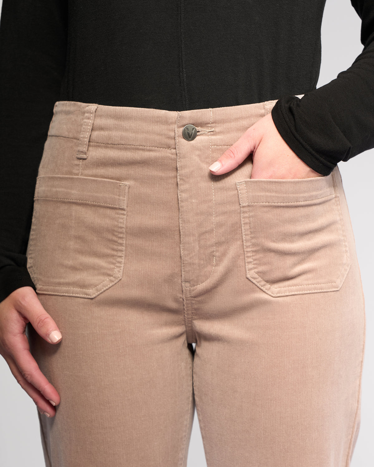 A person wears Vassalli's Daily Cord Pant - Twig, high-waisted beige corduroy pants with large patch pockets, paired with a black long-sleeve top. One hand is in a pocket, the other at their side.