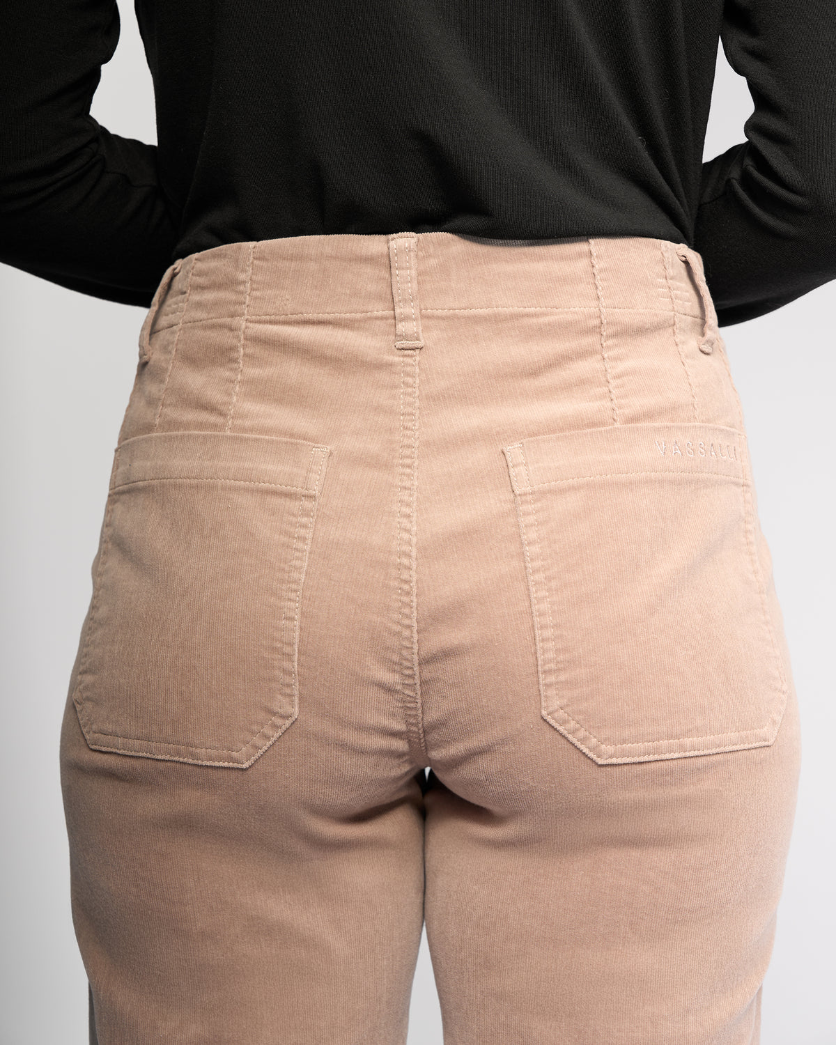 A person is seen from the waist down wearing Vassalli's Daily Cord Pant - Twig, a high-waisted, light beige corduroy style with ankle-grazer length, two large patch pockets, and a stitched brand label on the right pocket.