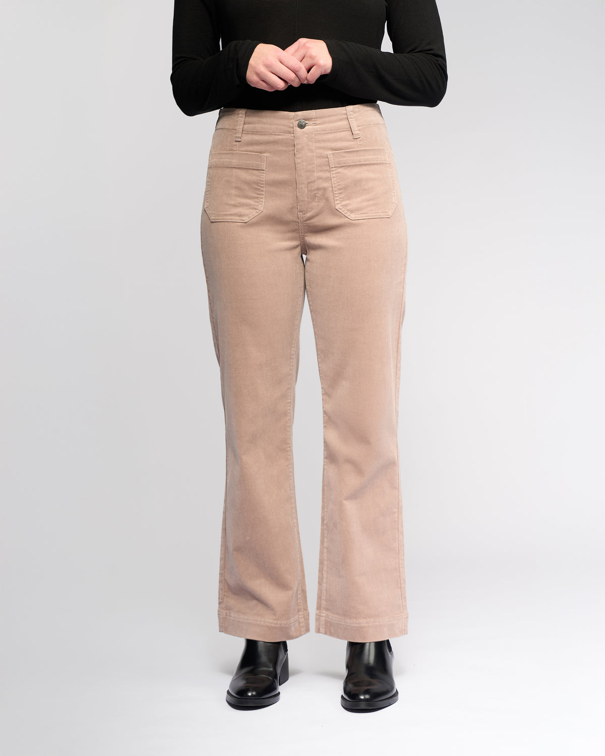 A person wears Vassalli's Daily Cord Pant - Twig in light beige with patch pockets, paired with a black long-sleeved top and black ankle boots, standing against a plain white background. Photo is cropped from the shoulders down.