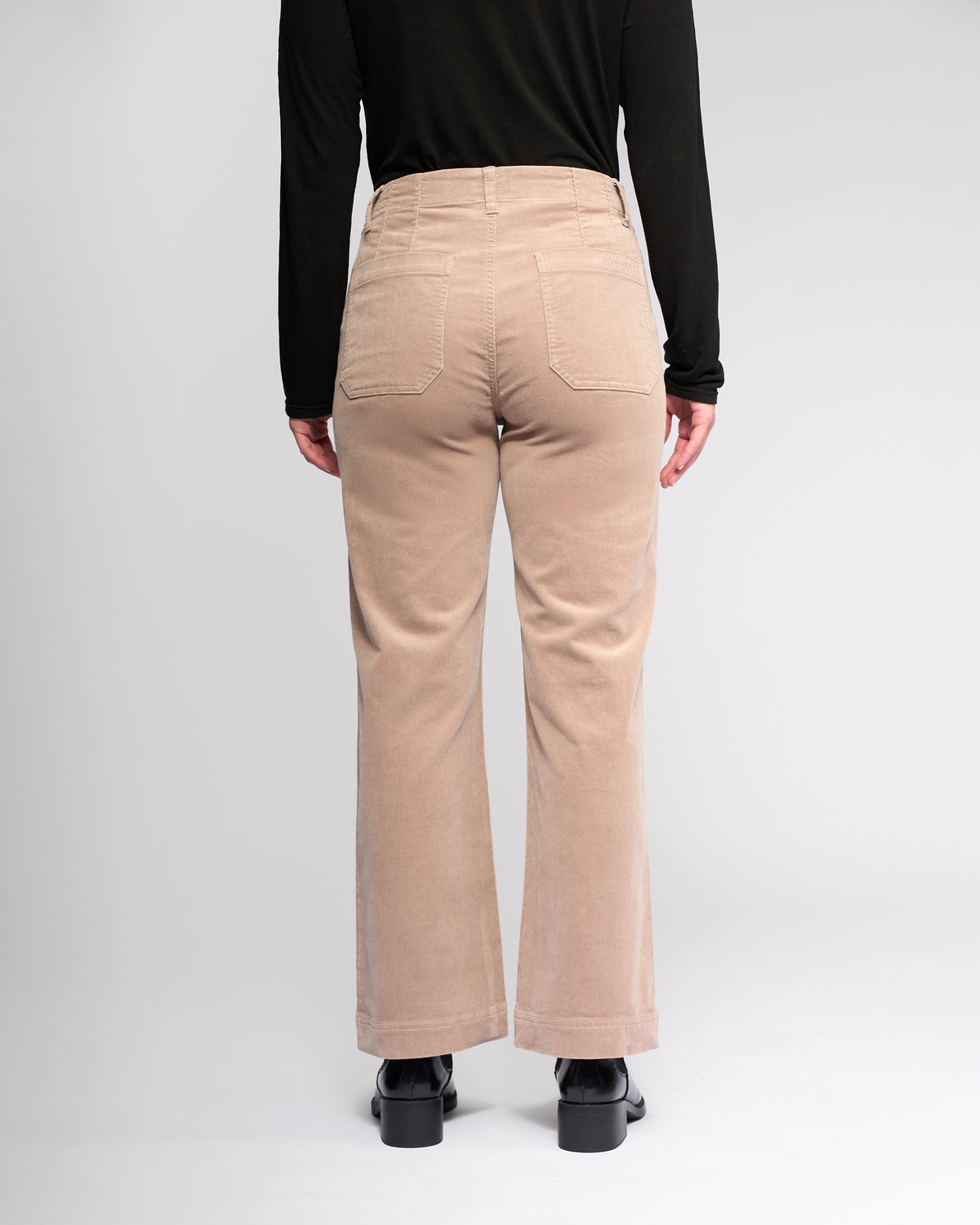 Someone wearing the Vassalli Daily Cord Pant in Twig, a black long-sleeve top, and black shoes stands facing away from the camera against a plain light background.