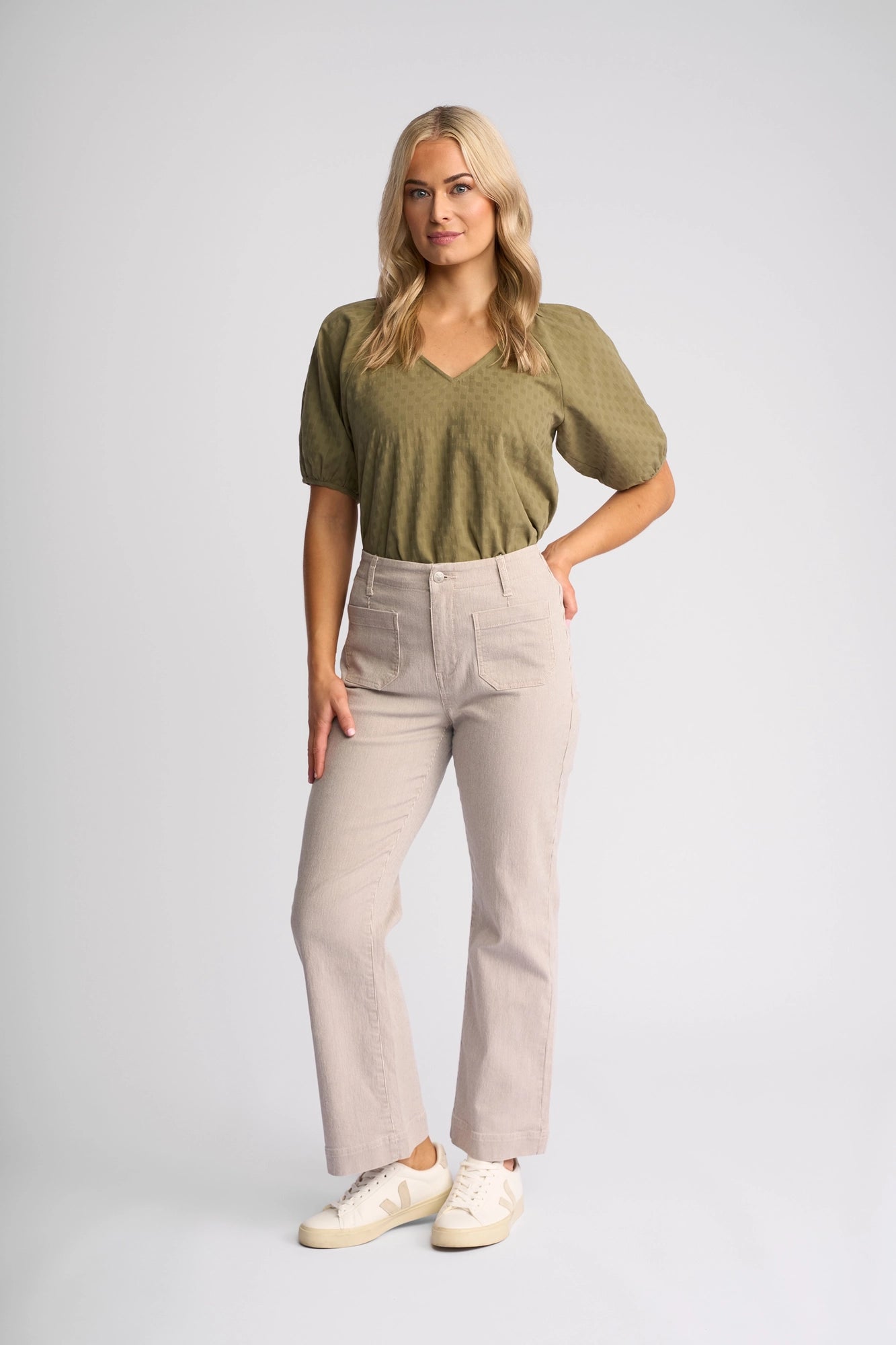 A woman with long blonde hair stands against a plain background, wearing an olive green short-sleeve top, the Vassalli Daily Jean in Ecru Stripe Denim, and white sneakers. She strikes a relaxed, confident pose with one hand on her hip.
